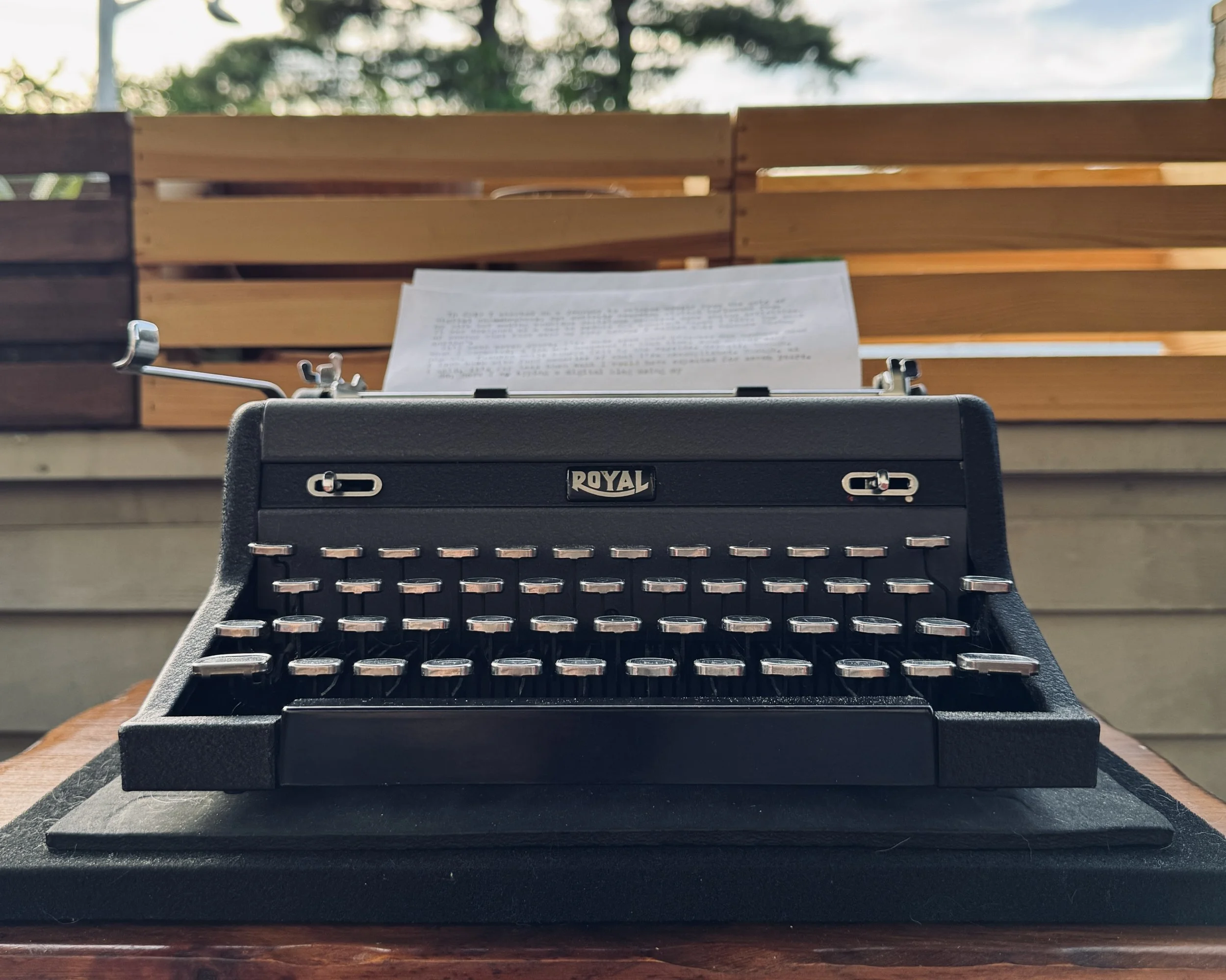 A black Royal typewriter sits on a wooden table outdoors, with a typed page loaded behind the keys. Warm evening light filters through the railing and trees in the background.