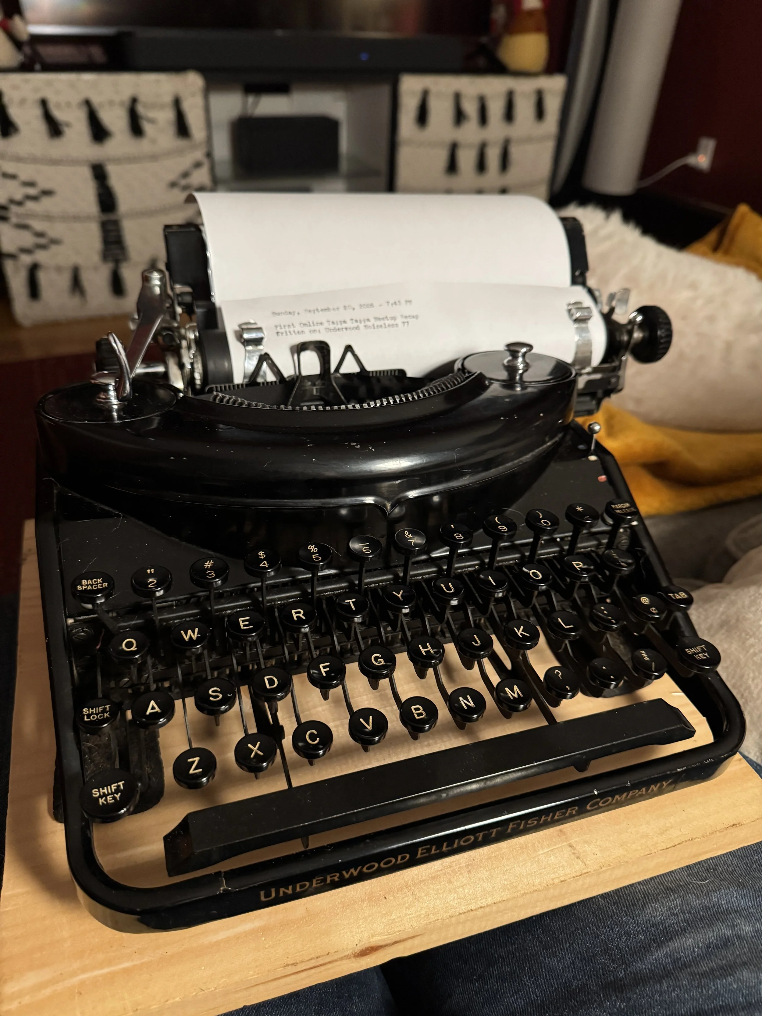 A black Underwood Noiseless 77 typewriter resting on a wooden lap desk, with a sheet of paper loaded into the platen. The machine sits in a cozy living room setting with blankets and a couch visible in the background.
