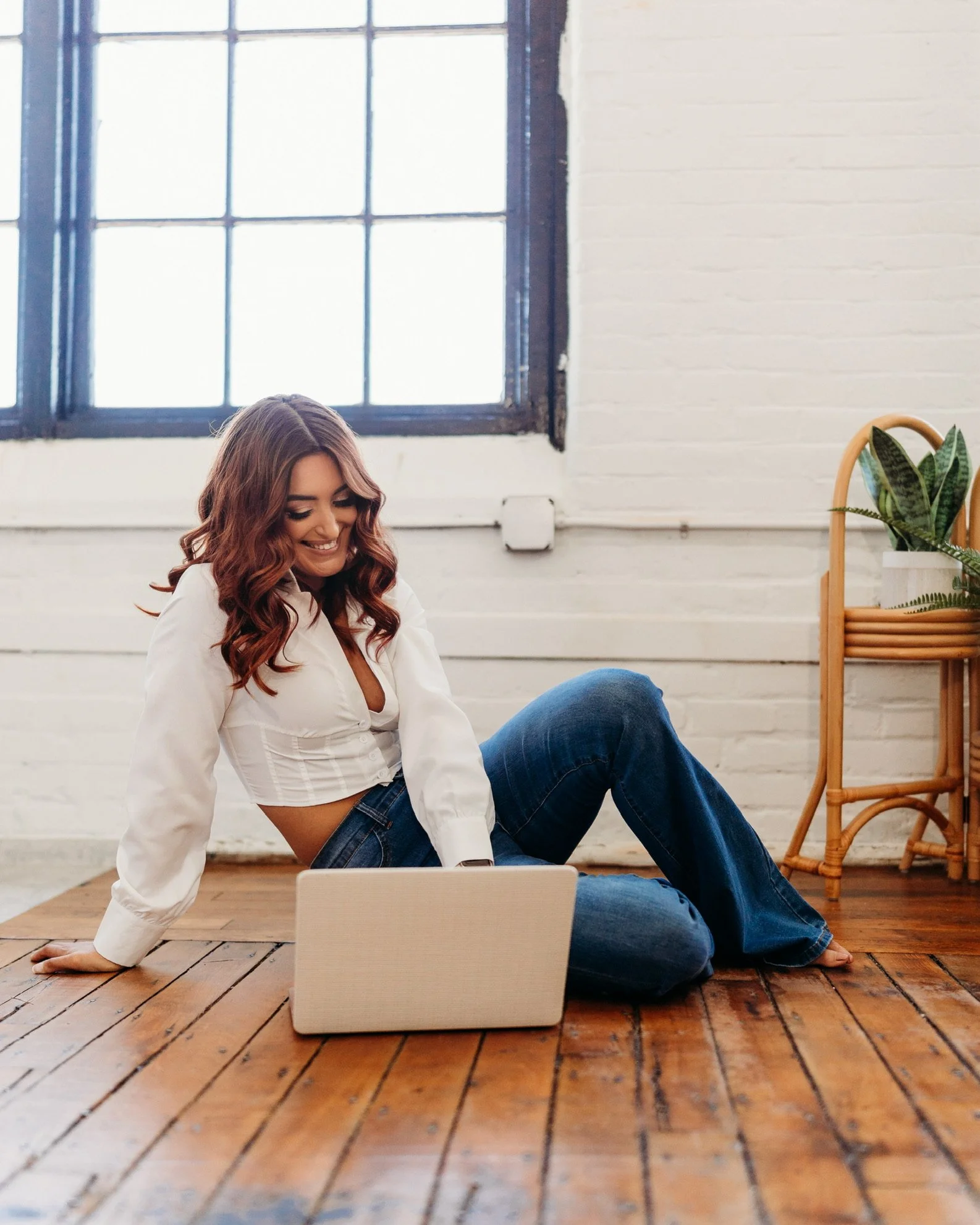 A woman with wavy brunette hair sitting on a wooden floor in a bright room, smiling at her laptop.