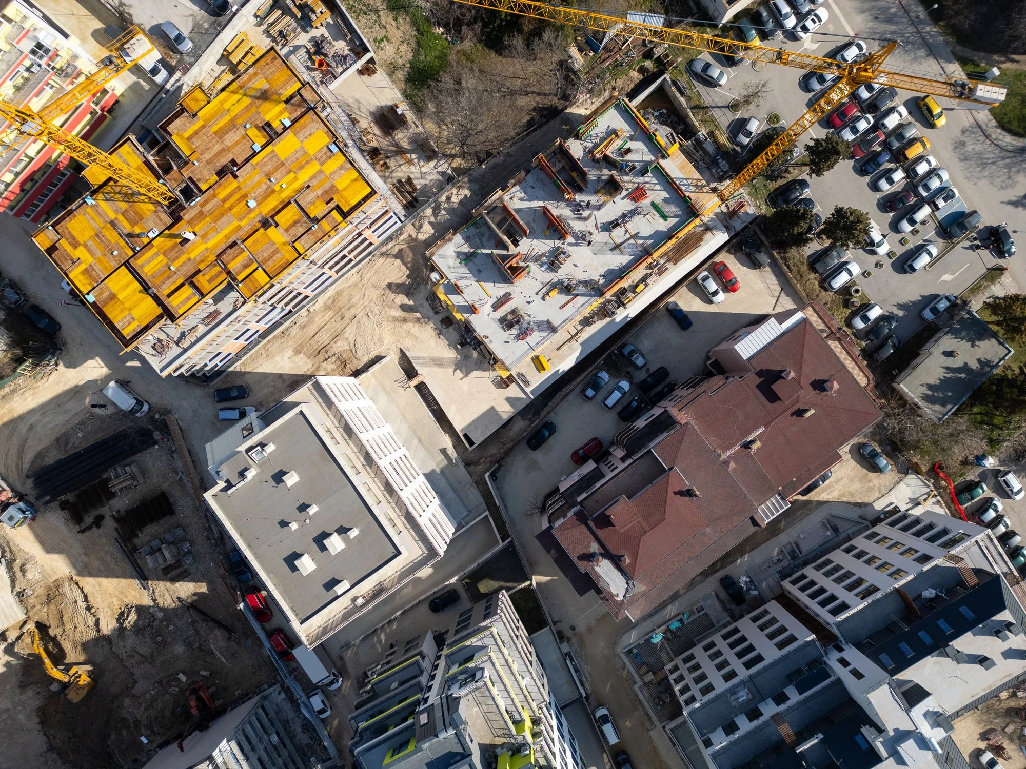 An aerial view of a construction site with a building in progress. The building has a yellow construction crane and partially completed floors with some in wood framing and others with concrete slabs. Surrounding the site are parked cars and nearby buildings.