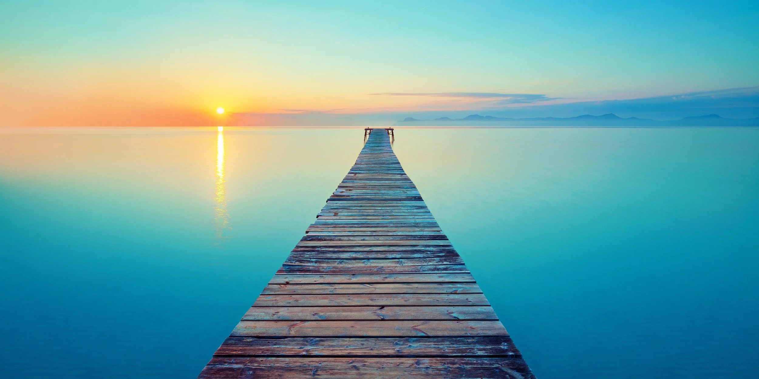 Wooden pier extending into a calm body of water during sunset, with mountains in the distance and a clear sky.