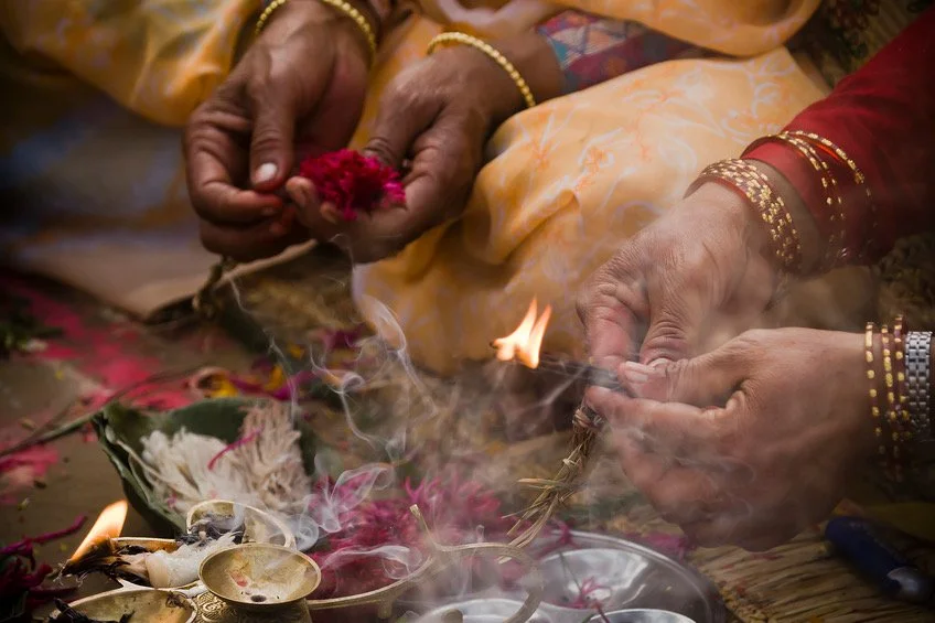 Two people performing a traditional ritual, holding lit incense sticks over a decorated altar with flowers and incense.
