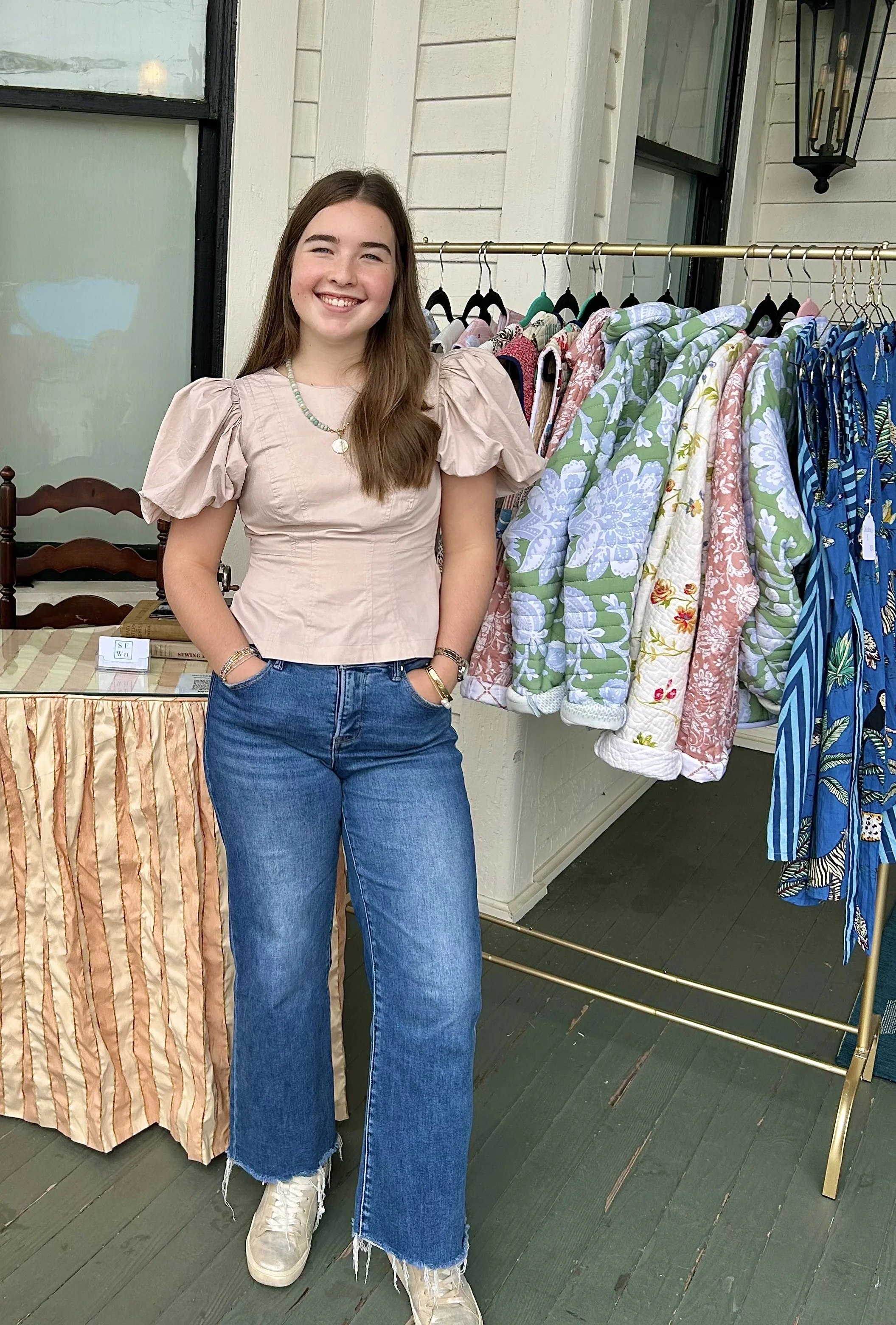 A young woman with long brown hair, wearing a beige puff-sleeve top, blue jeans, and sneakers, standing next to a rack of colorful patterned jackets and a table behind her at an outdoor market or store.