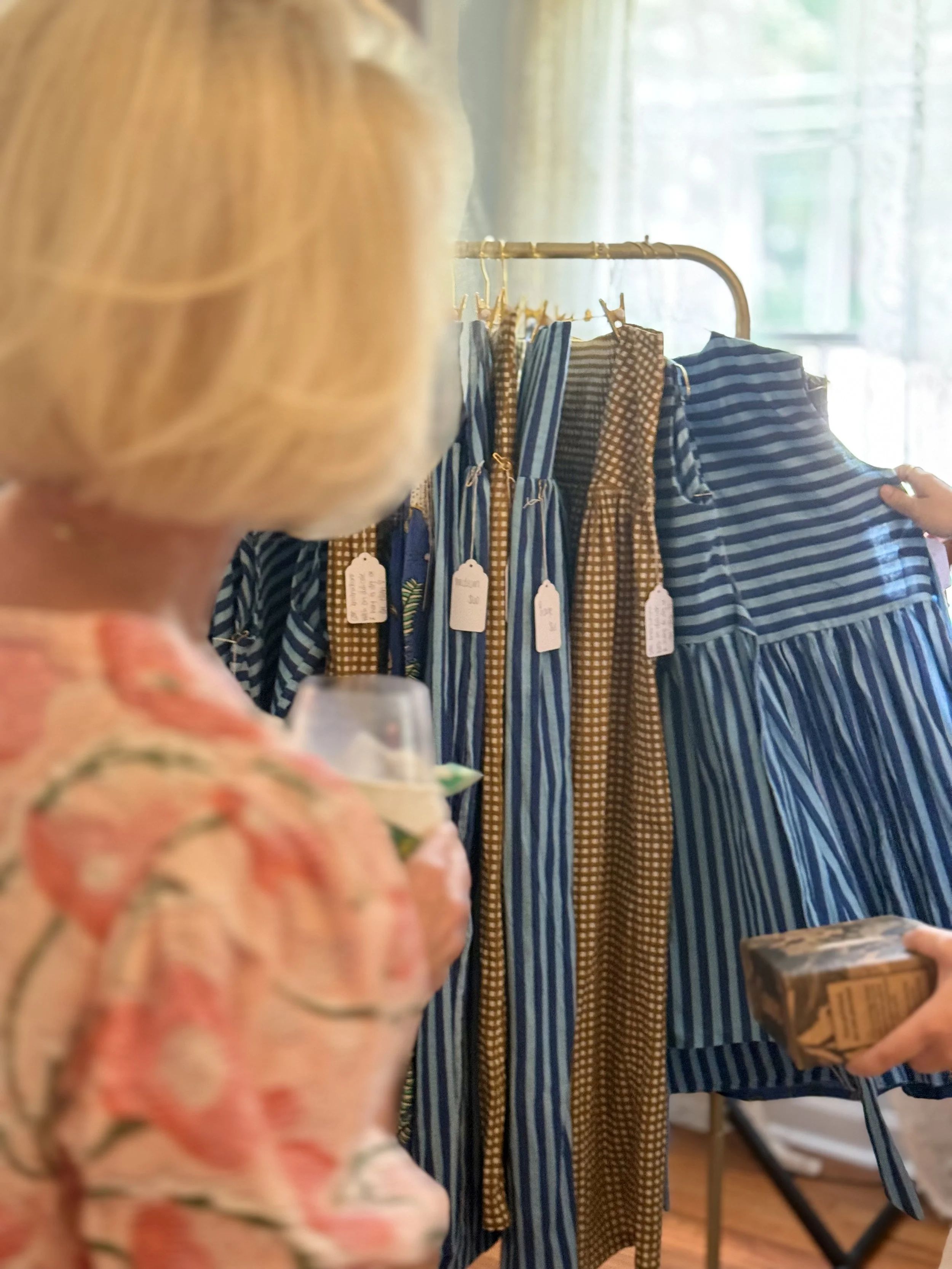 A person with blonde hair looking at blue and brown striped and checkered dresses on a clothing rack in a sunlit room.