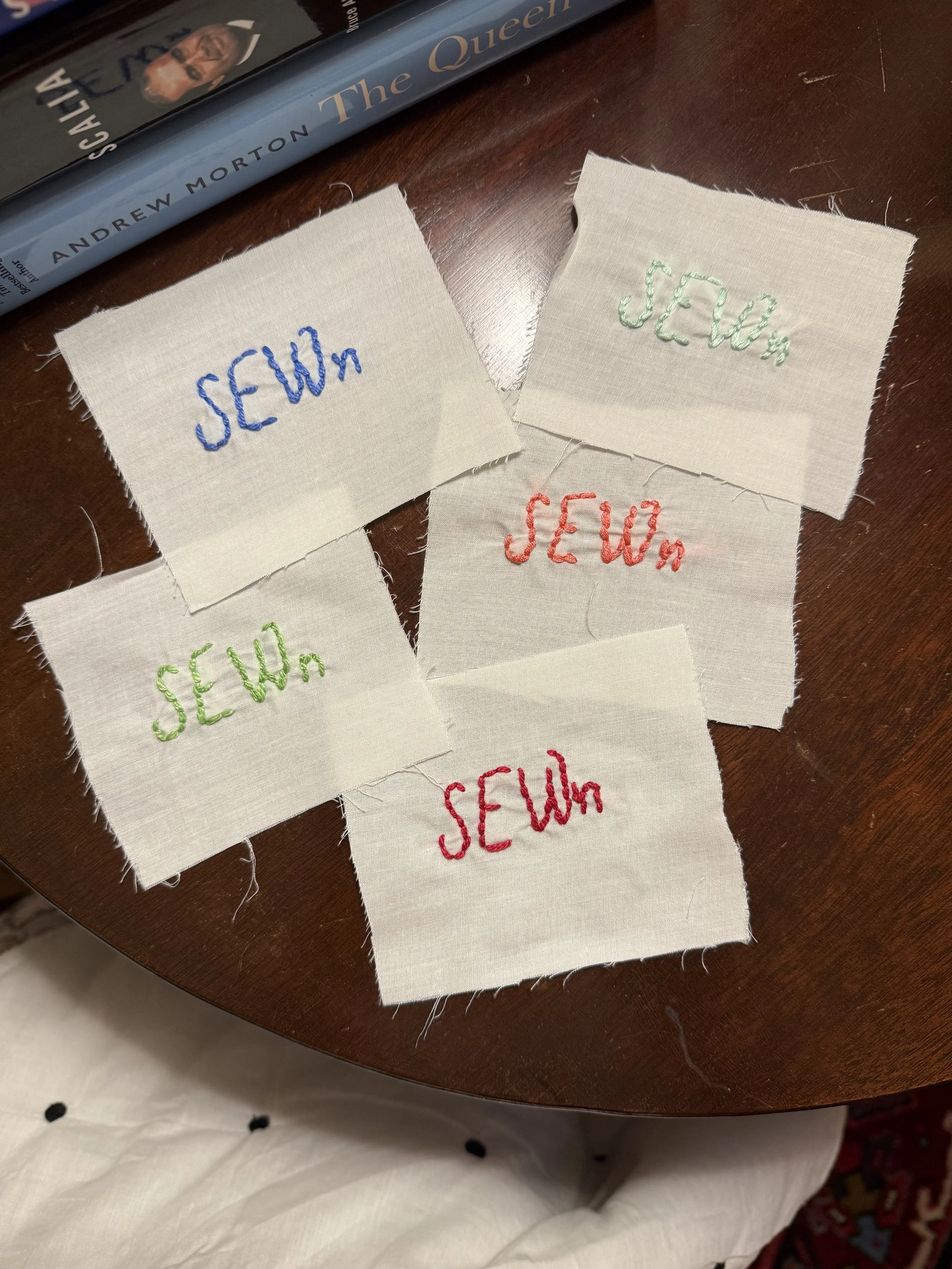 Several small fabric squares with the embroidered initials 'SEWn' in different colors, placed on a wooden table. A book titled 'The Queen' by Andrew Morton is partially visible in the background.