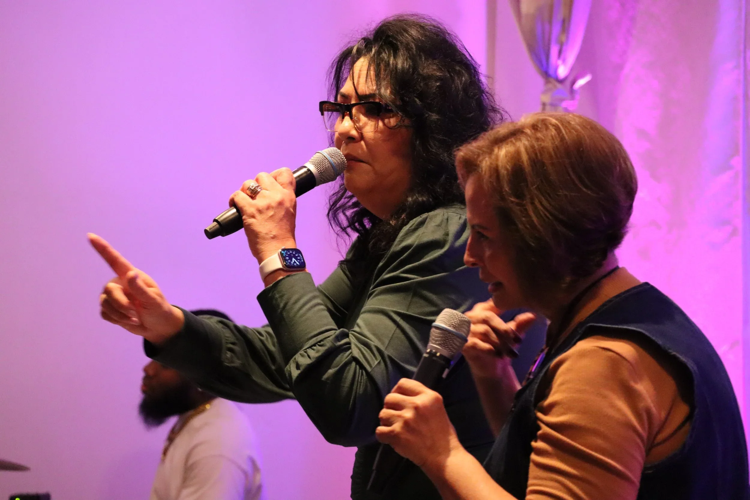 Two women speaking into microphones at an indoor event with purple lighting. One woman has dark curly hair, glasses, and is wearing a gray jacket, gesturing with her left hand. The other woman has short hair and is wearing a brown shirt with a sleeve