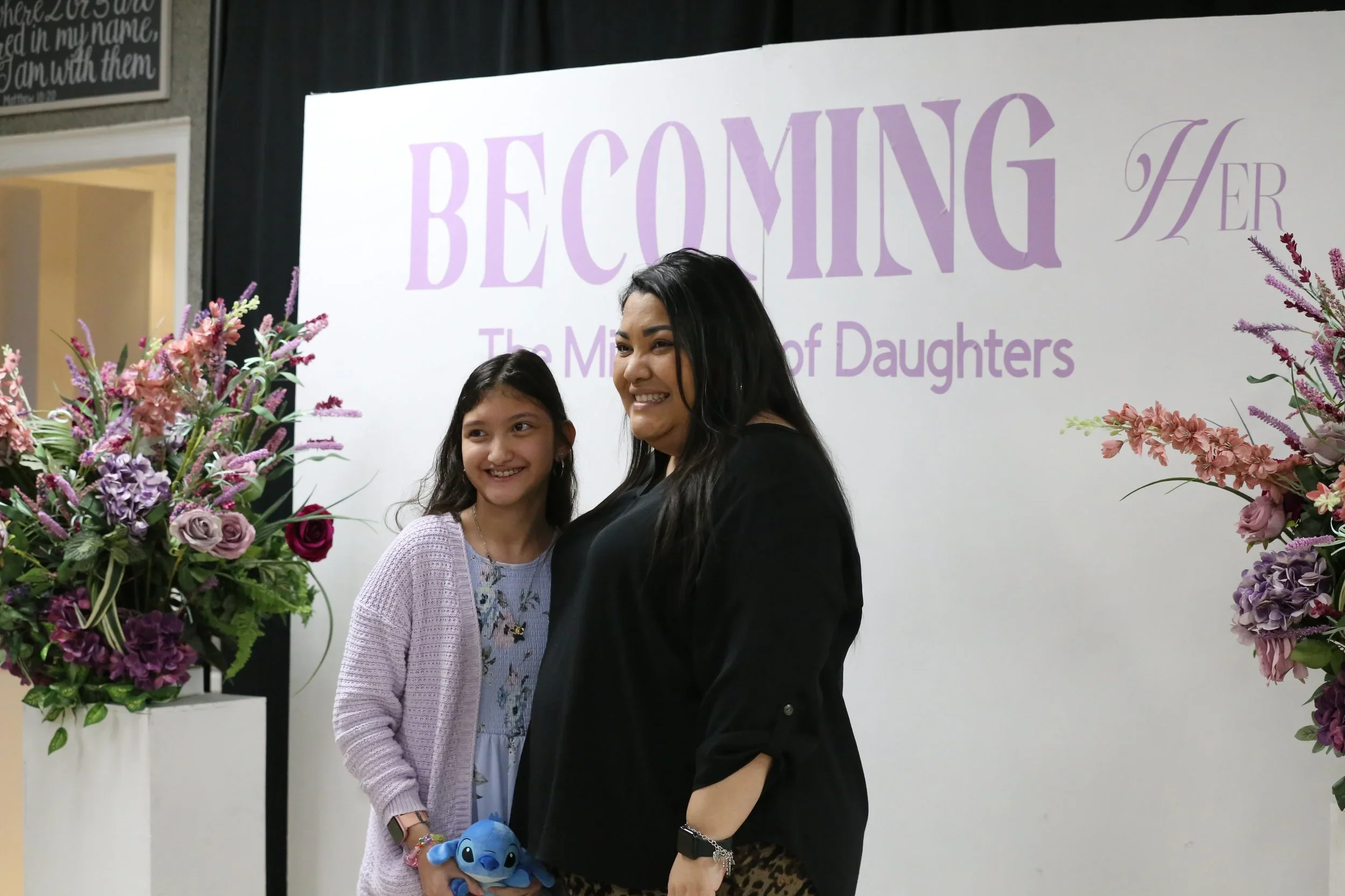 A woman and a girl smiling and standing next to each other at an event with a backdrop that reads 'BECOMING HER The Making of Daughters', surrounded by floral arrangements.