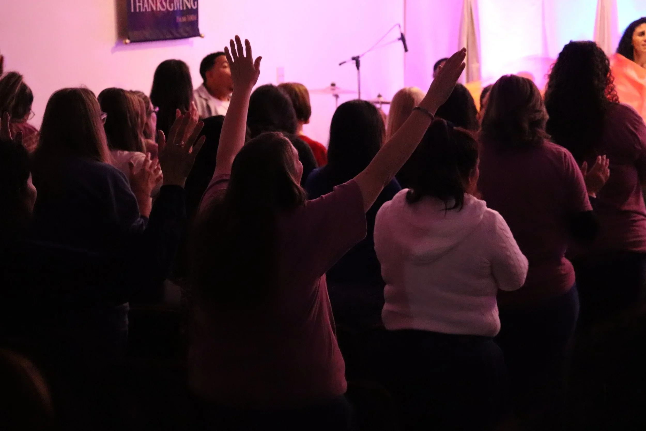 People sitting in a darkened room with pink lighting, raising their hands, during a gathering or event, with a stage and microphone in the background.