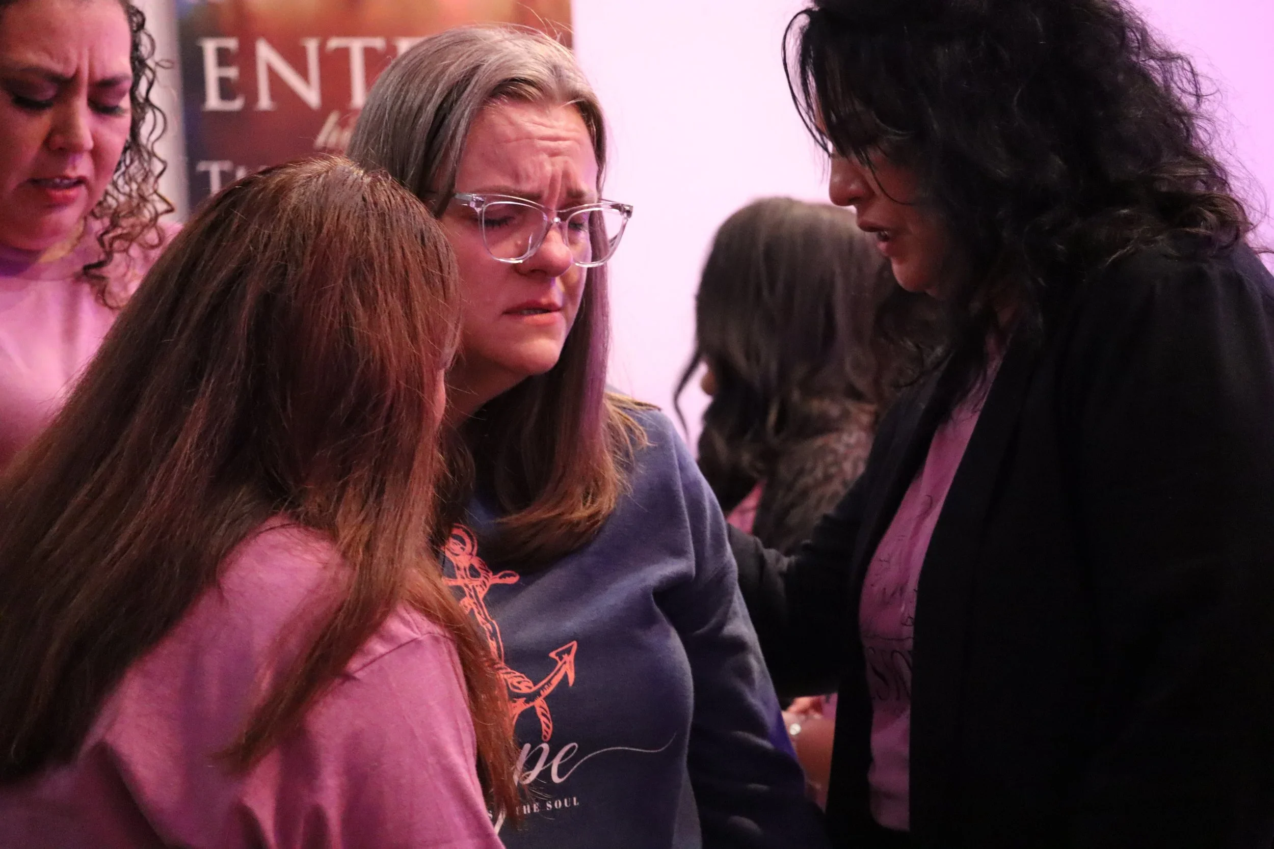 Four women are gathered closely together with their eyes closed, participating in a prayer or moment of reflection. The woman in the foreground has long red hair and is wearing a pink shirt. The woman next to her has long, straight brown hair, glasse