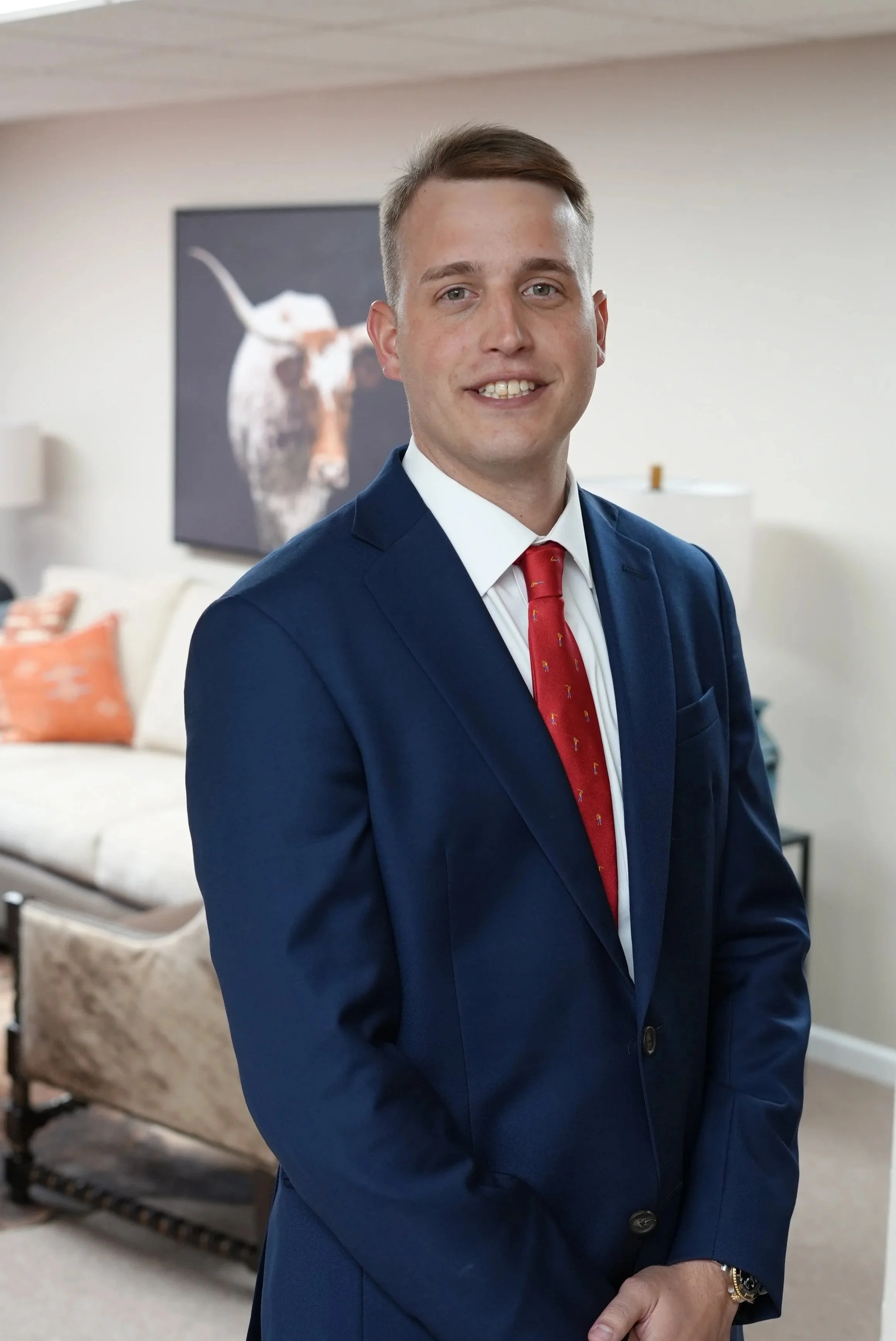 A young man in a navy blue suit and red tie standing indoors, smiling at the camera with a living room setting in the background, including a white sofa, orange cushions, and a piece of artwork featuring a cow.