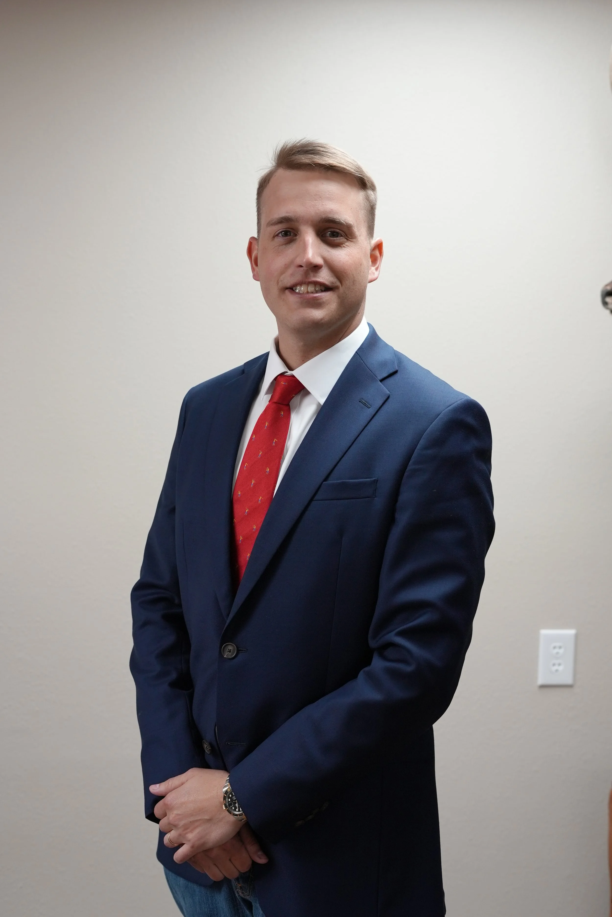 A young man with light skin, short light brown hair, wearing a navy blazer, white shirt, and red tie with small yellow patterns, standing against a plain beige wall. He is smiling and has his hands clasped in front of him. He is wearing a watch on his left wrist.