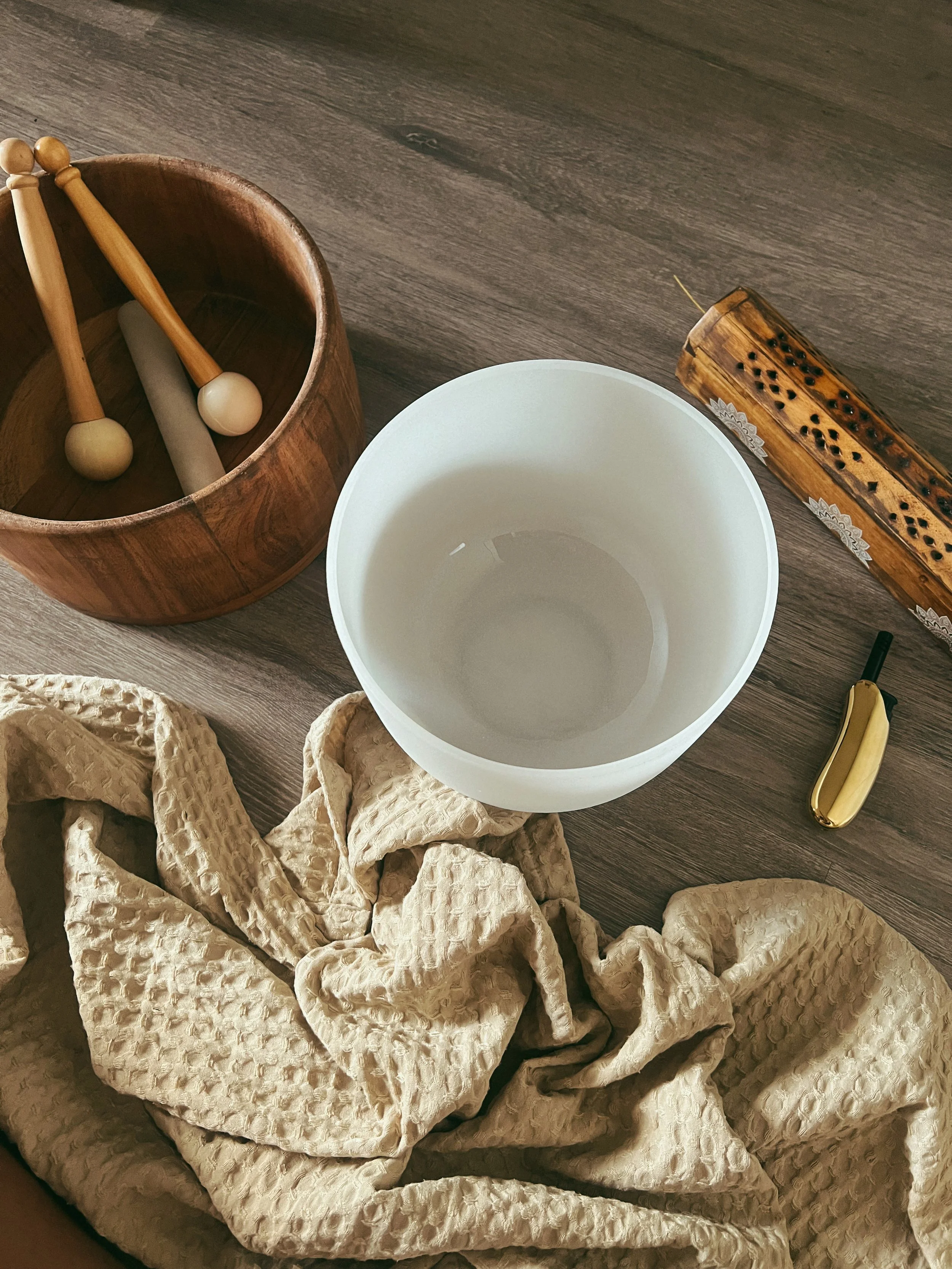 An arrangement of therapy musical instruments and objects on a wooden floor, including a wooden box with mallets, a sound bowl, a wooden grated instrument, a banana, and a crumpled textured cloth.
