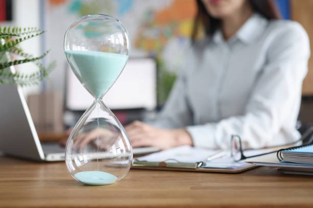 An hourglass with light blue sand on a wooden desk, with a therapist doing supervision virtually in the background.