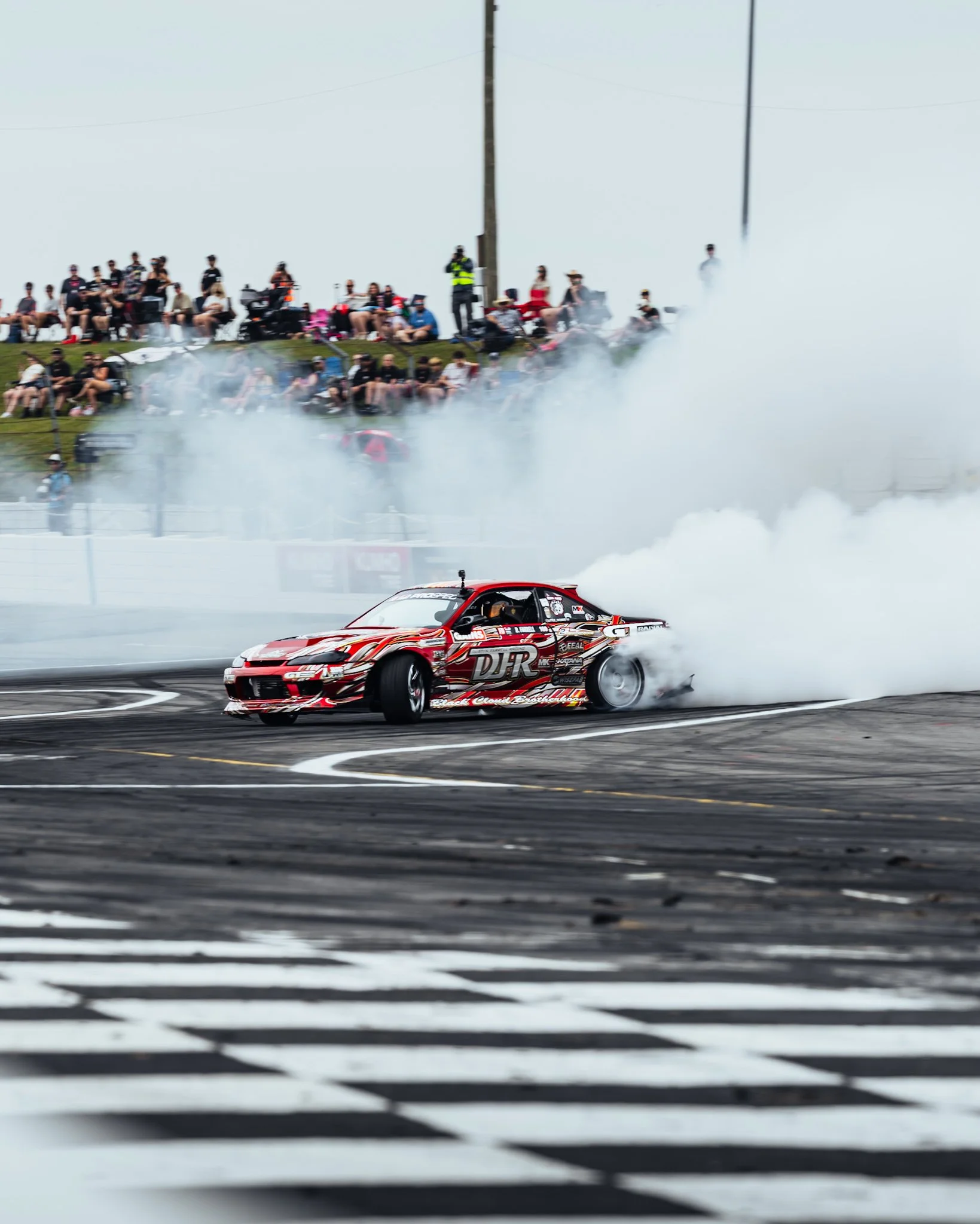 A race car performing a burnout on a racing track, creating tire smoke with spectators and photographers watching from a grassy hill above the track. Formula Drift Indy