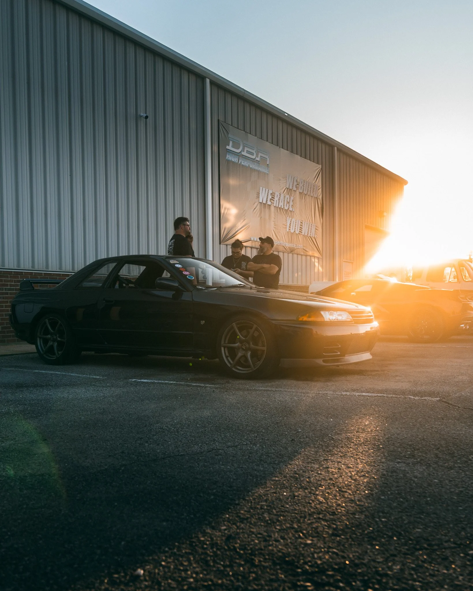 Three men standing outside a black sports car, which is parked in front of a large metal building with a banner. The sun is setting, casting a warm glow, with additional cars parked nearby.