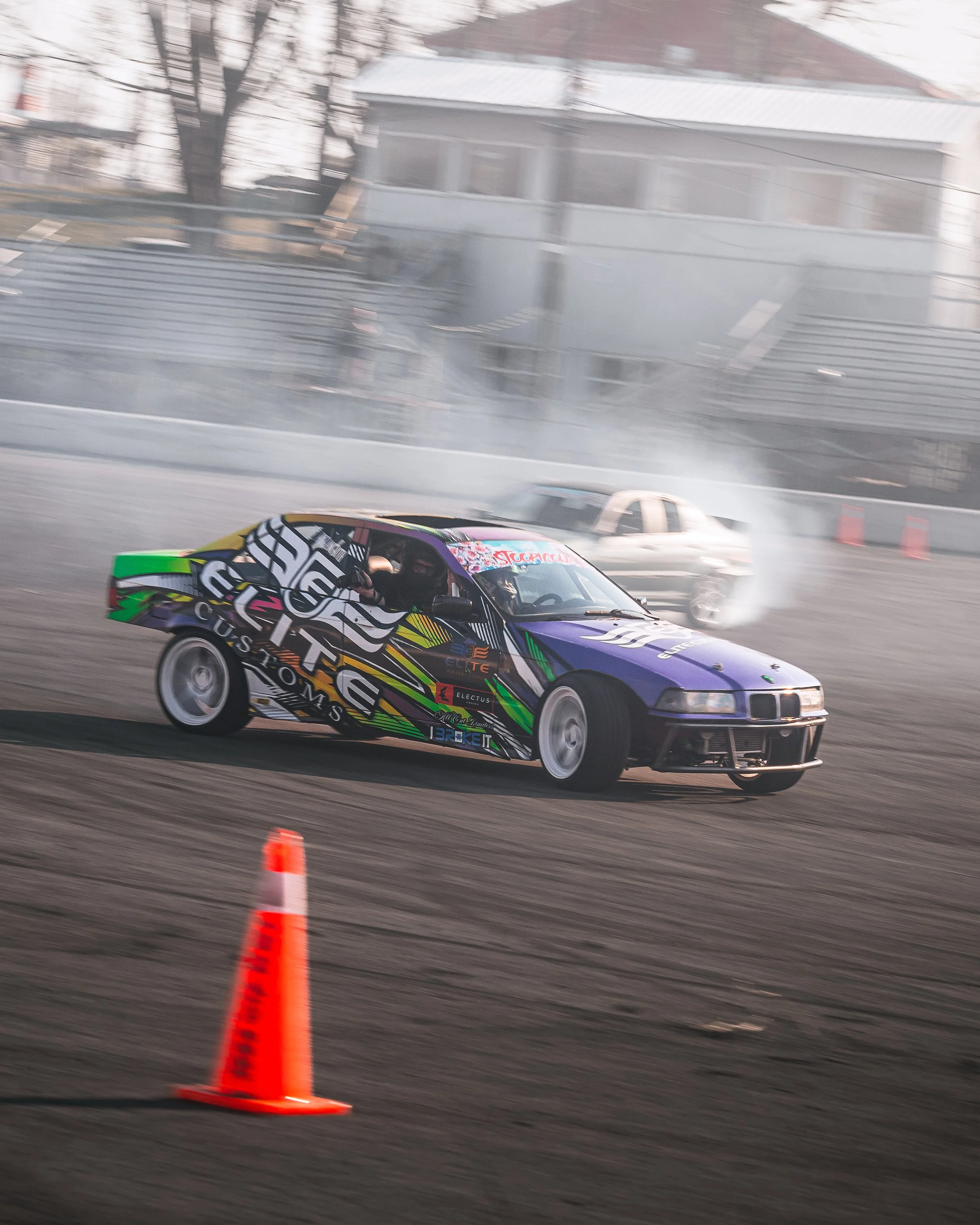 A colorful race car drifting on a track with smoke and tire marks, orange traffic cone in foreground, and another vehicle in the background.