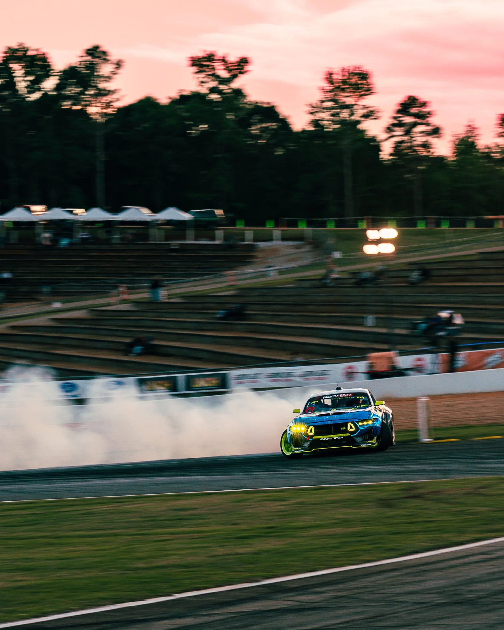 A blue and black race car drifting on a race track, creating white smoke from the tires, during sunset with a pink sky and trees in the background.