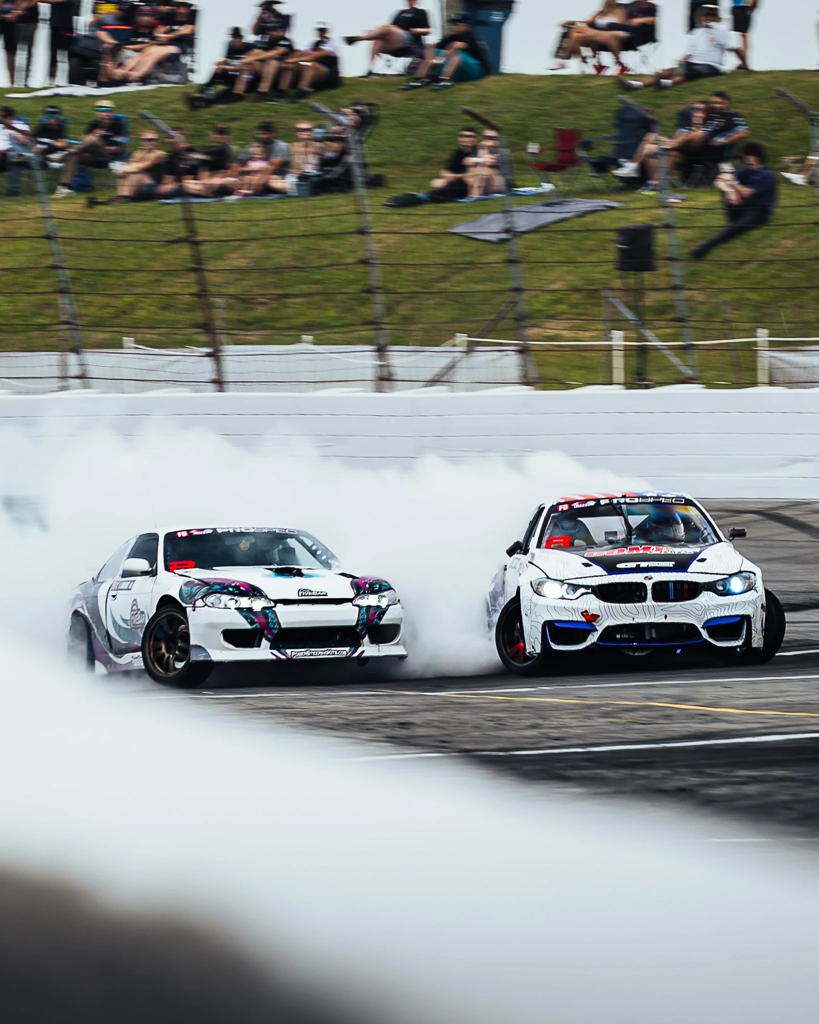 Two race cars speeding on a race track, with skid marks on the pavement, surrounded by a white barrier, while spectators watch from a grassy hillside behind a wire fence.