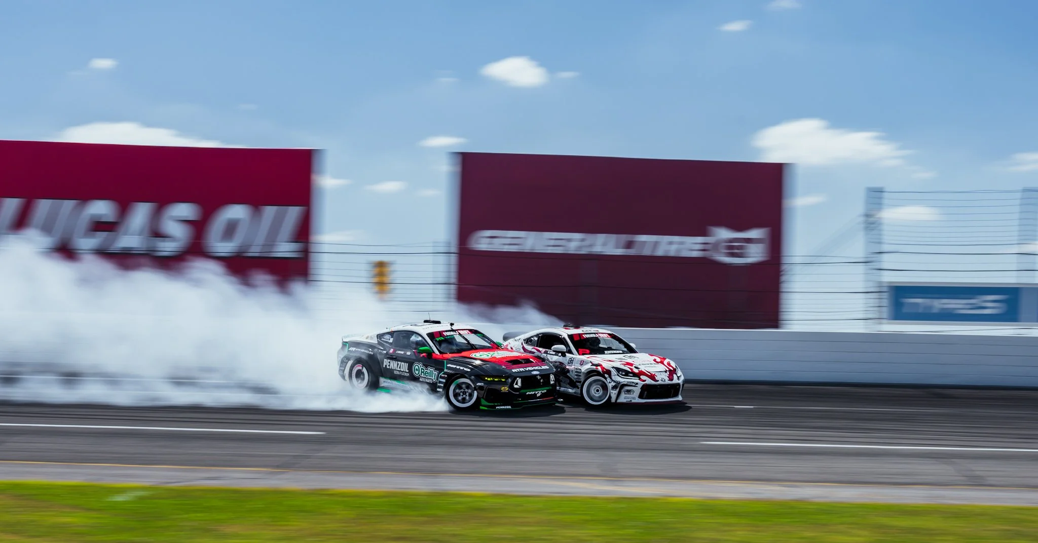 Two race cars drifting on a race track, creating smoke behind them, with advertisements and a fence in the background. Formula Drift Indy