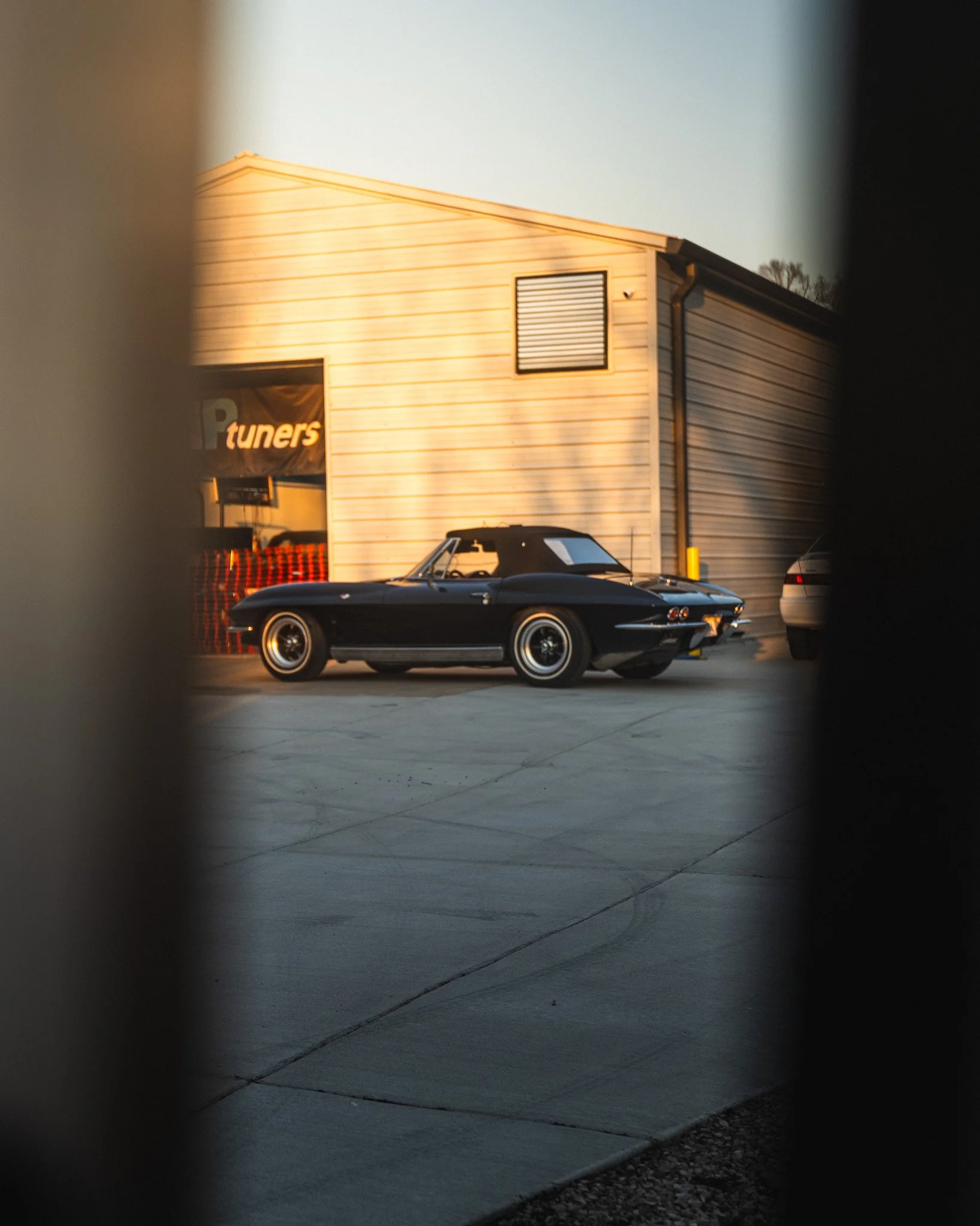 A black vintage convertible car parked outside a beige building during sunset, viewed through a gap between objects.
