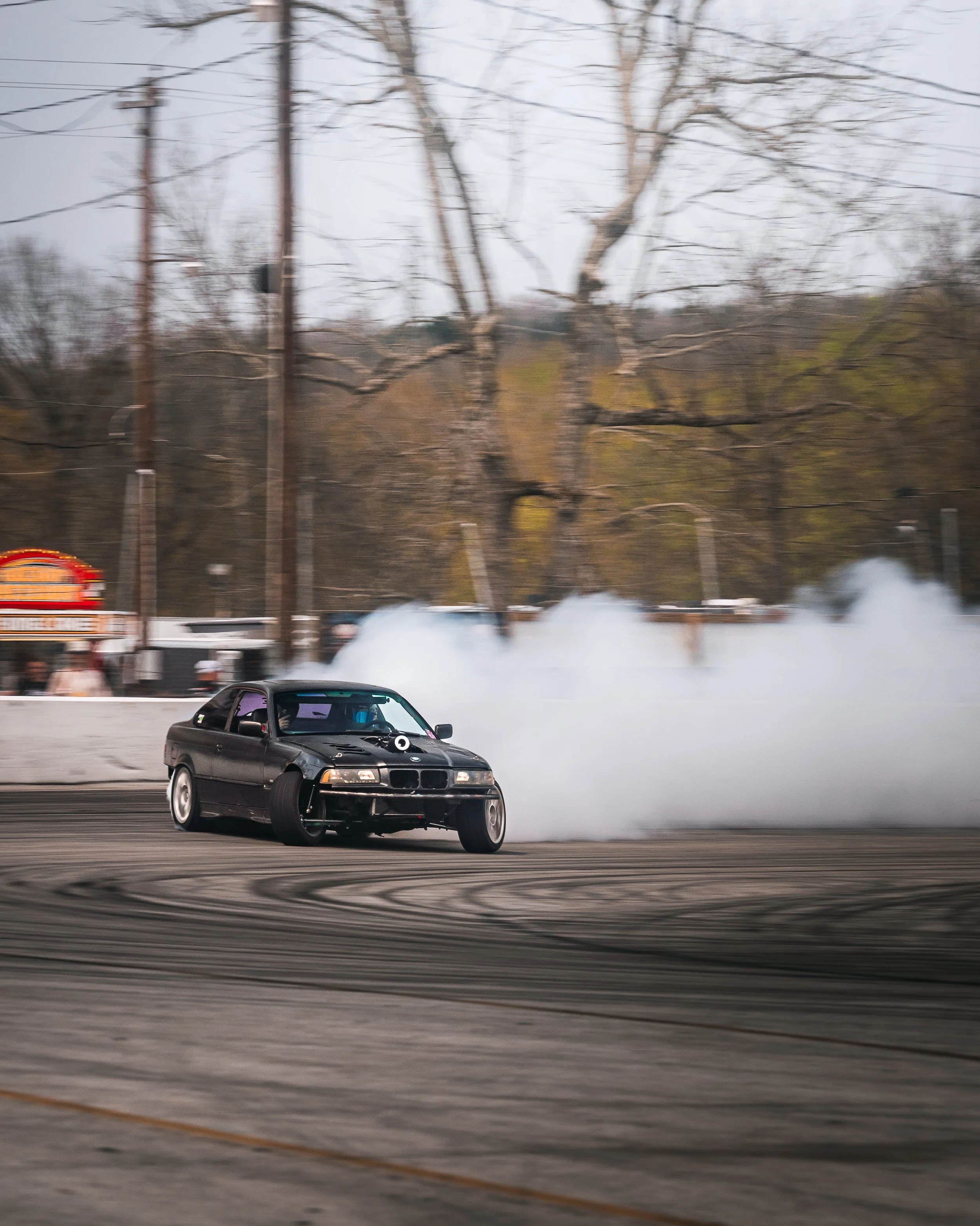 A black car performing a drift around a corner, creating a large cloud of smoke on an outdoor racing track, with leafless trees and utility poles in the background.