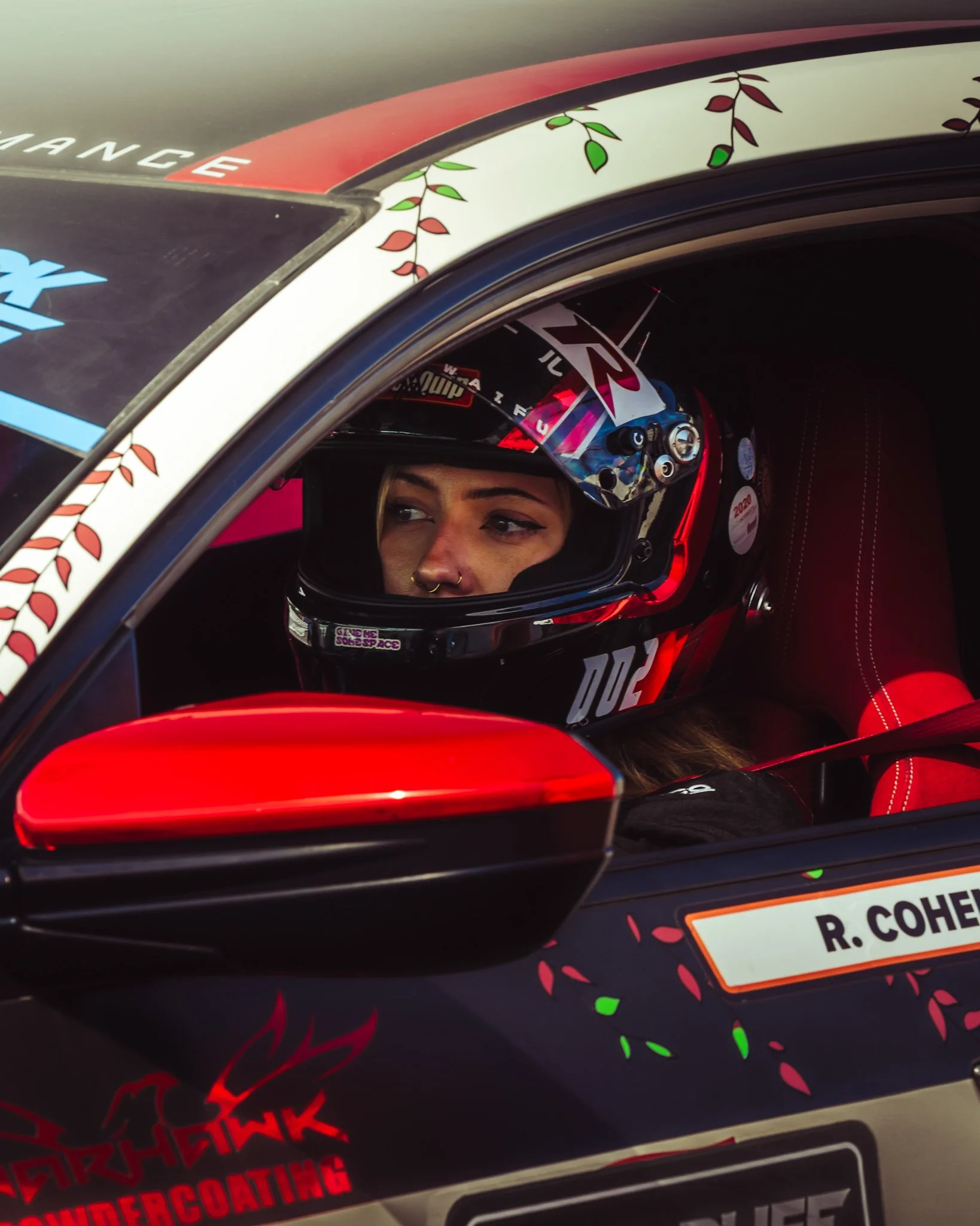 Close-up of race car window featuring decorative painted leaves and an athlete wearing a racing helmet with the name R. Cohen on the driver's nameplate.