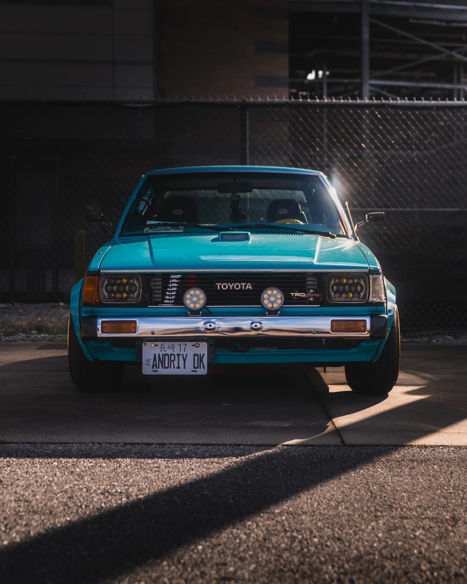 A vintage teal Toyota car with custom front lights and Japanese license plates parked in front of a chain-link fence with a dark building in the background.