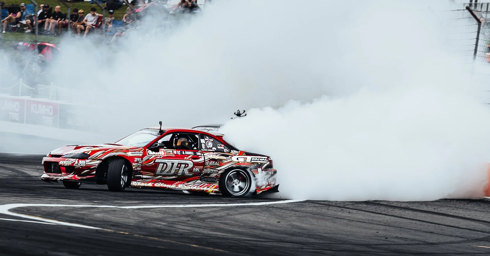 A drifting race car producing smoke as it takes a turn on a race track during a motorsport event, with spectators watching from the sidelines. Formula Drift Indy