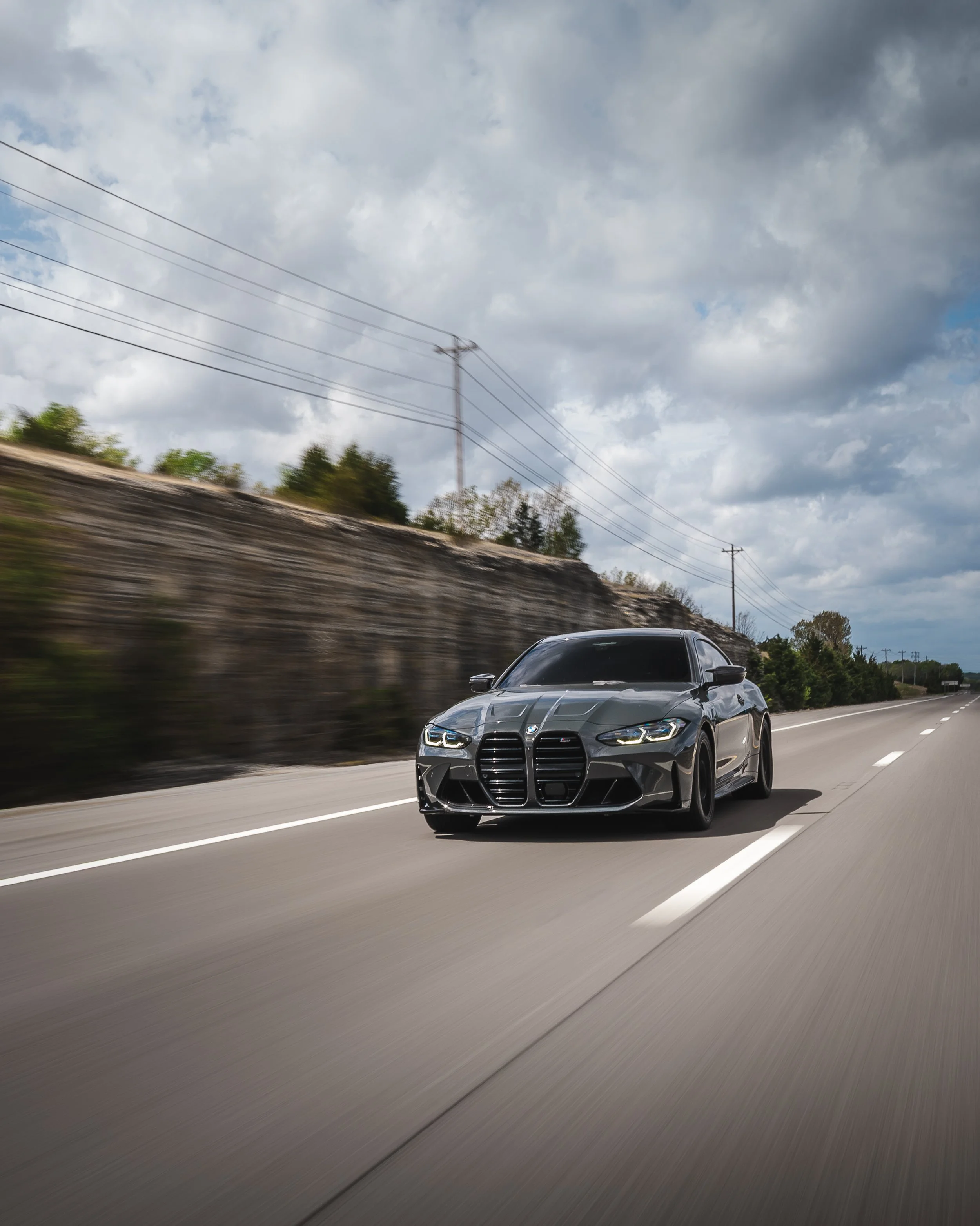Gray BMW car driving on a highway with a cloudy sky overhead and power lines in the background.
