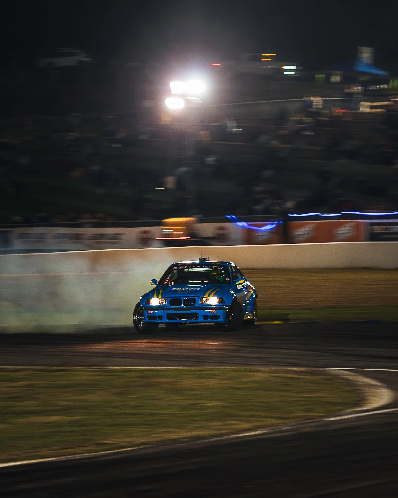 A blue race car with yellow and black accents driving on a race track at night, with bright headlights and blurred background of other vehicles and track barriers.