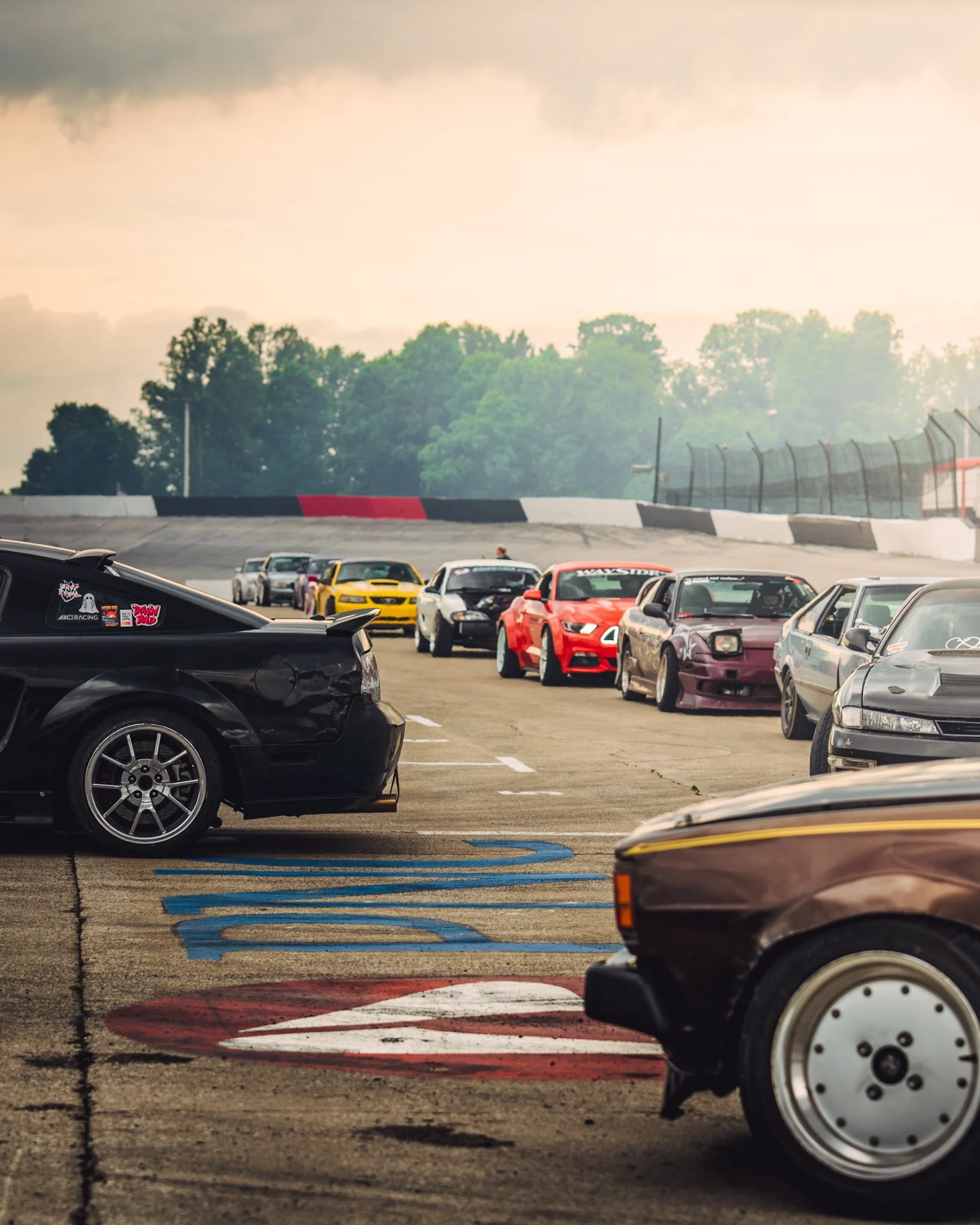 A parking lot at a racetrack filled with various sports cars, including a black car on the left with stickers, a yellow car, a red car, and several others, with tire barriers and a fence in the background, trees, and a cloudy sky.
