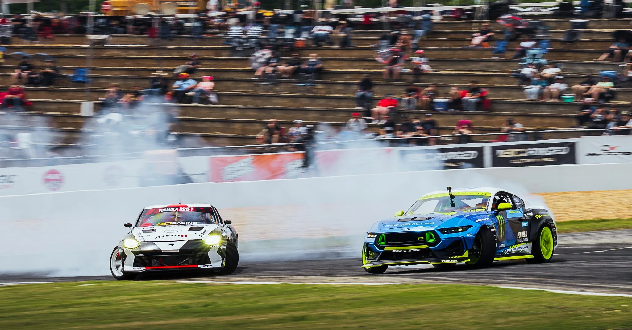 Two race cars drifting on a track with smoke and tire marks, while spectators sit on bleachers in the background.