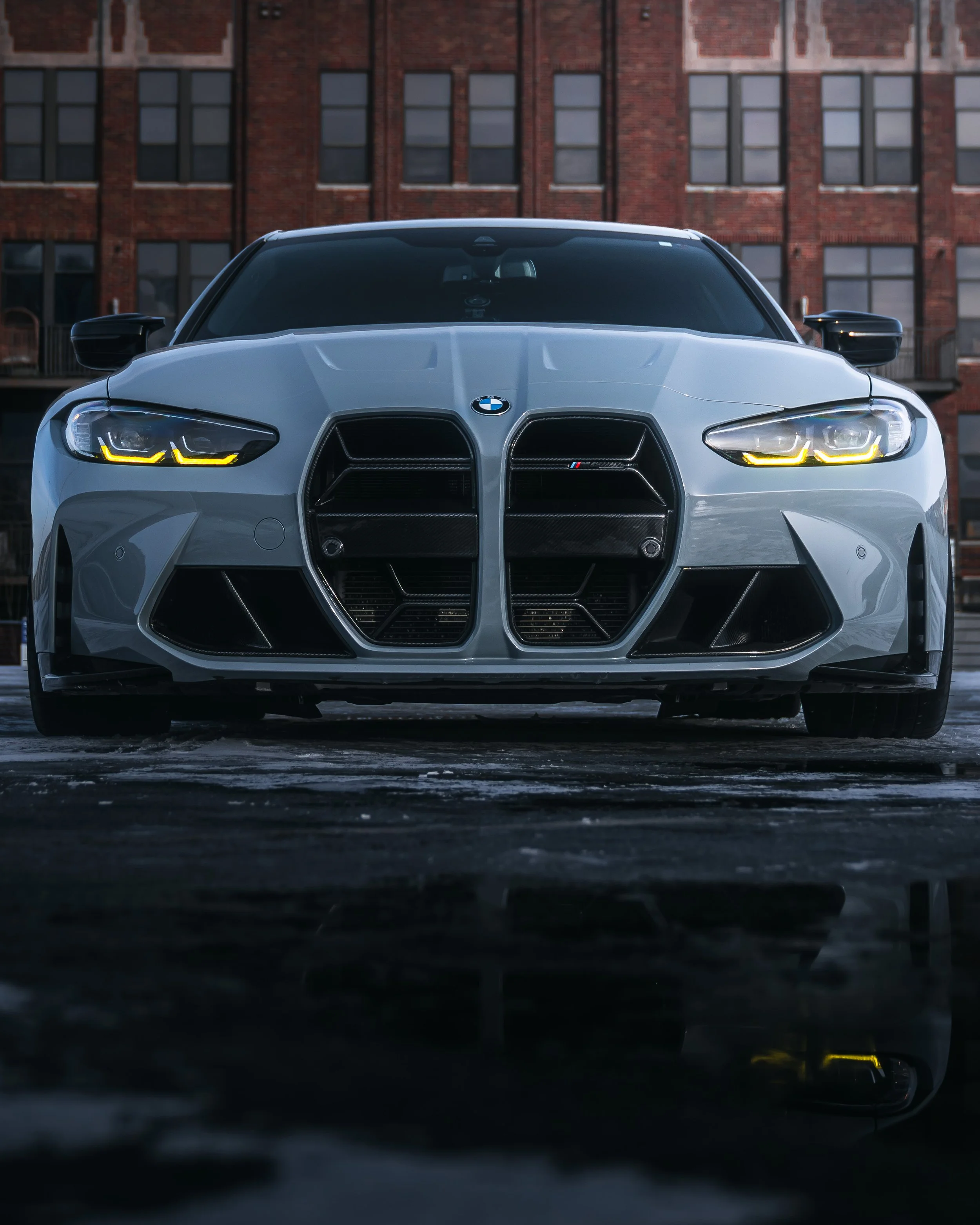 Front view of a white BMW sports car with illuminated headlights, parked on an urban street with brick buildings in the background.