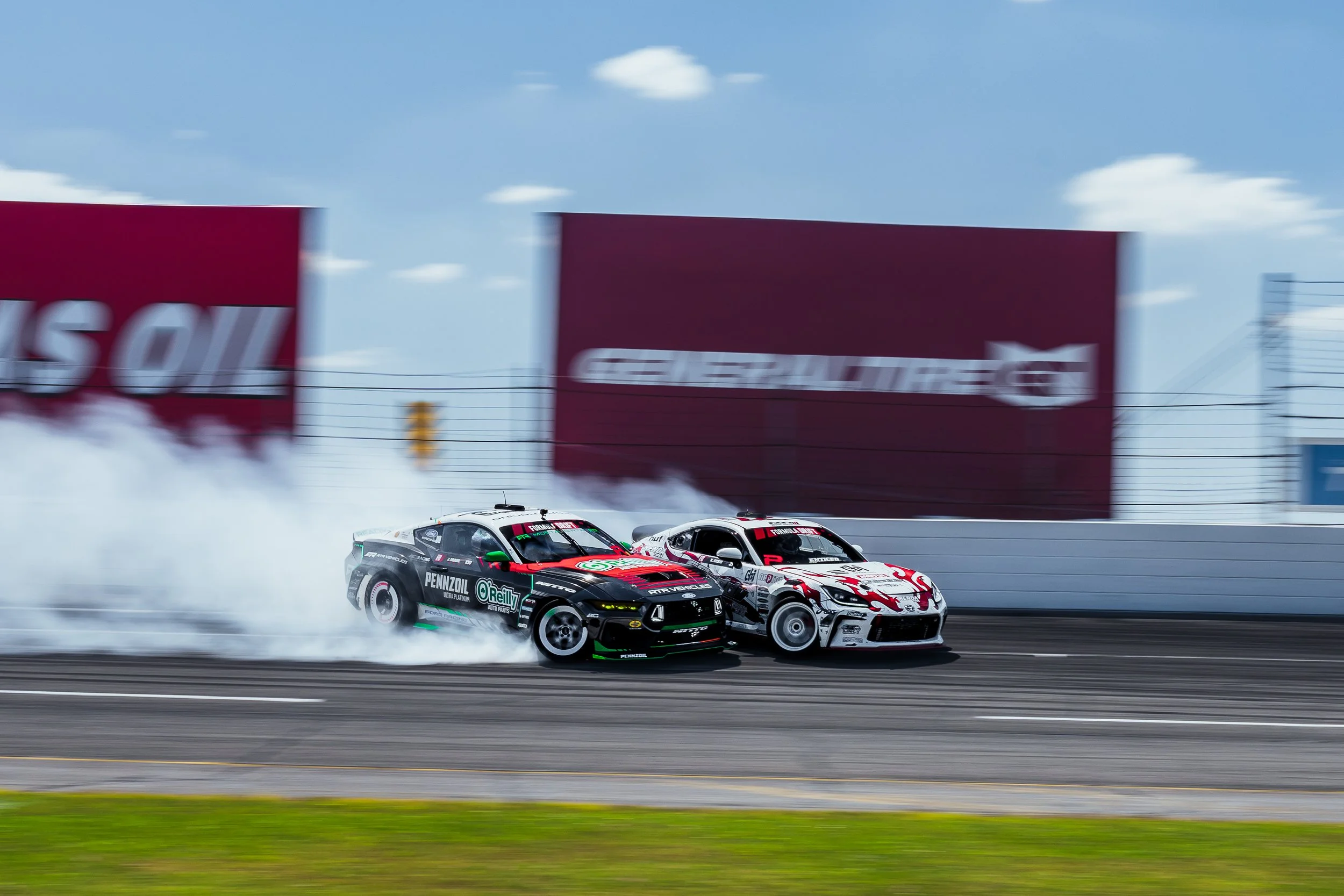 Two race cars speeding around a track, kicking up snow, with a blue sky and clouds in the background. One car is predominantly black with green and red accents, the other is predominantly white with red accents. Formula Drift Indy