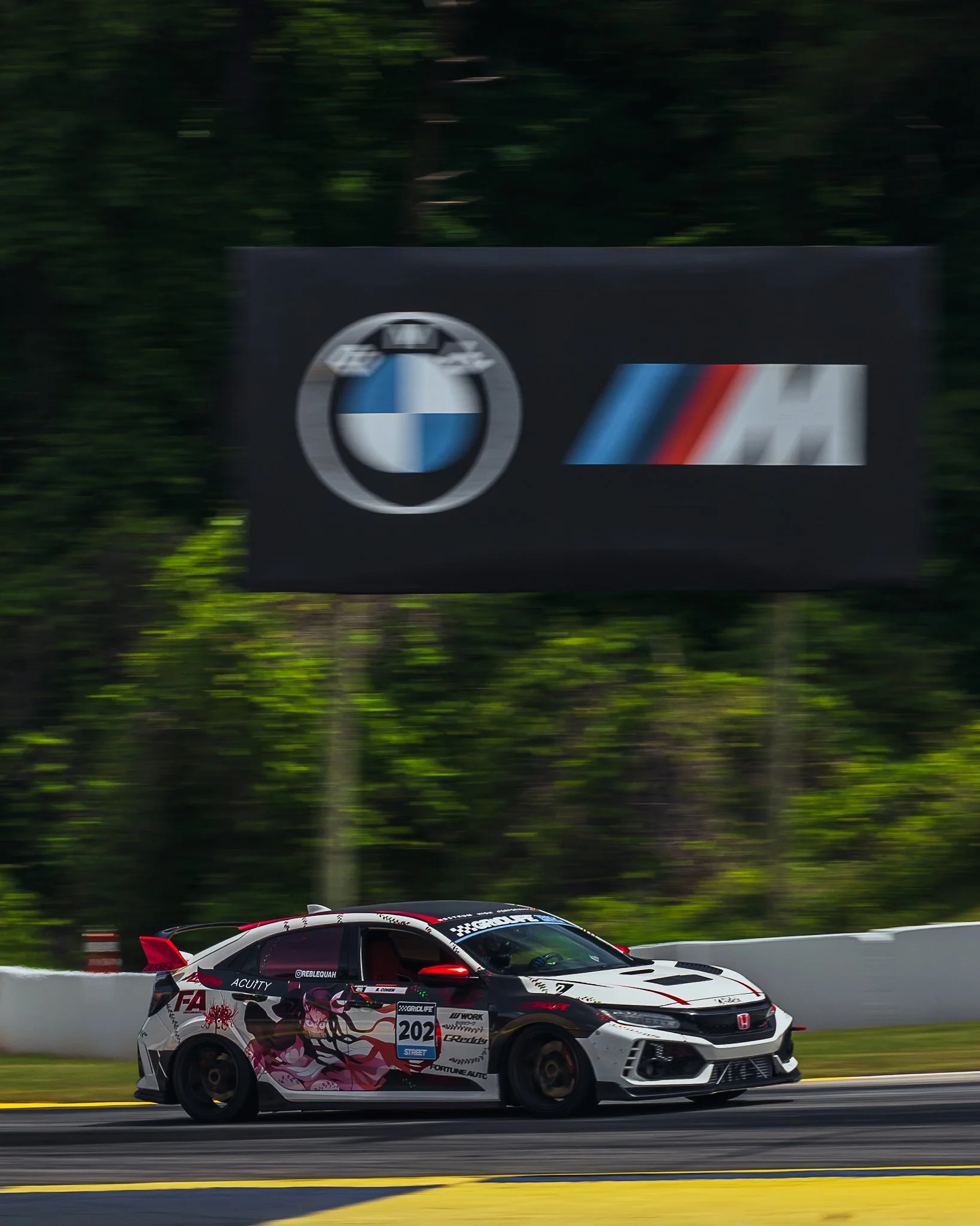 A race car with anime-style graphics driving on a track with a forest background.