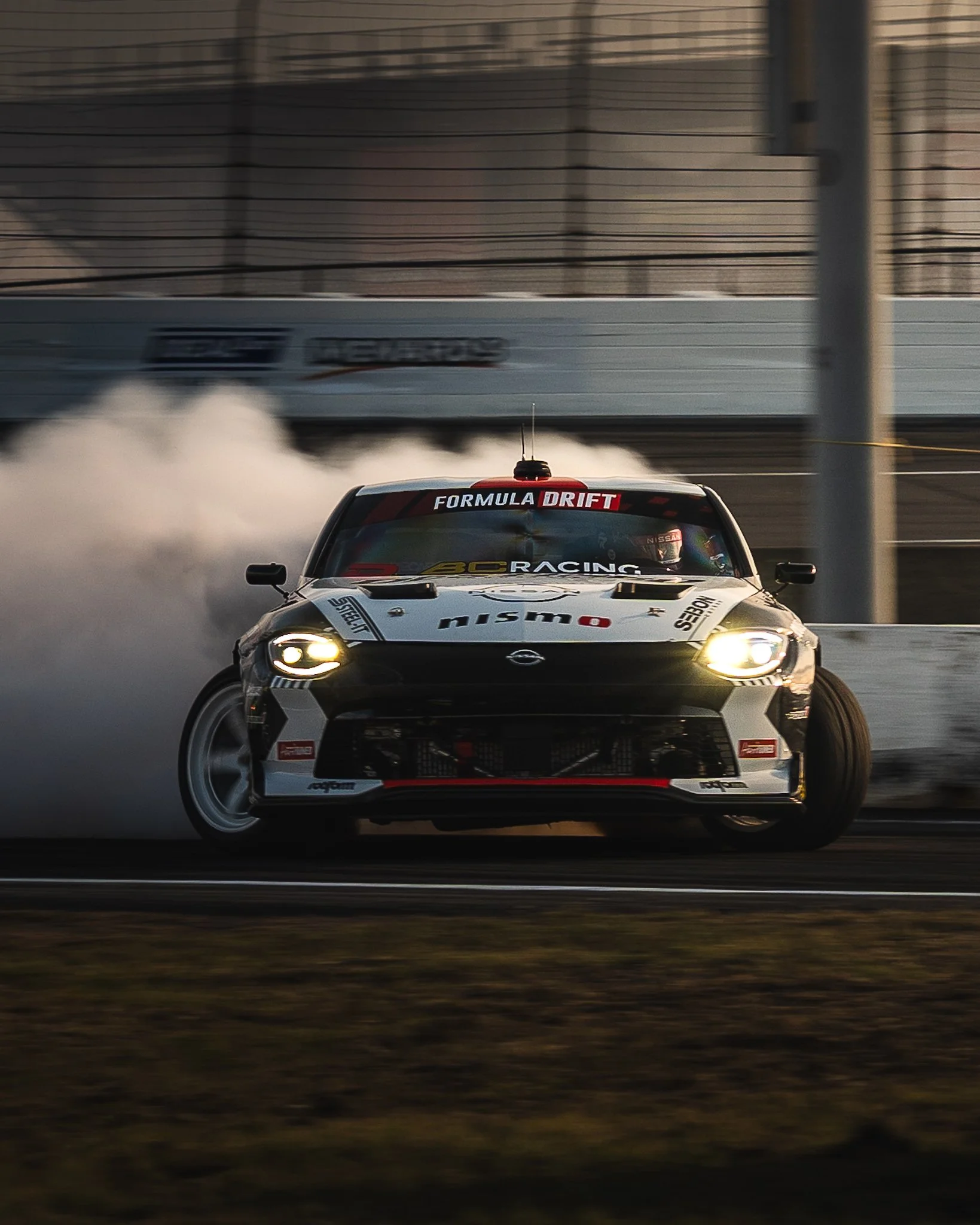A race car moving on a track with smoke trailing from the tires, with a fence and advertising banners in the background. Chris Forsberg Formula Drift Indy