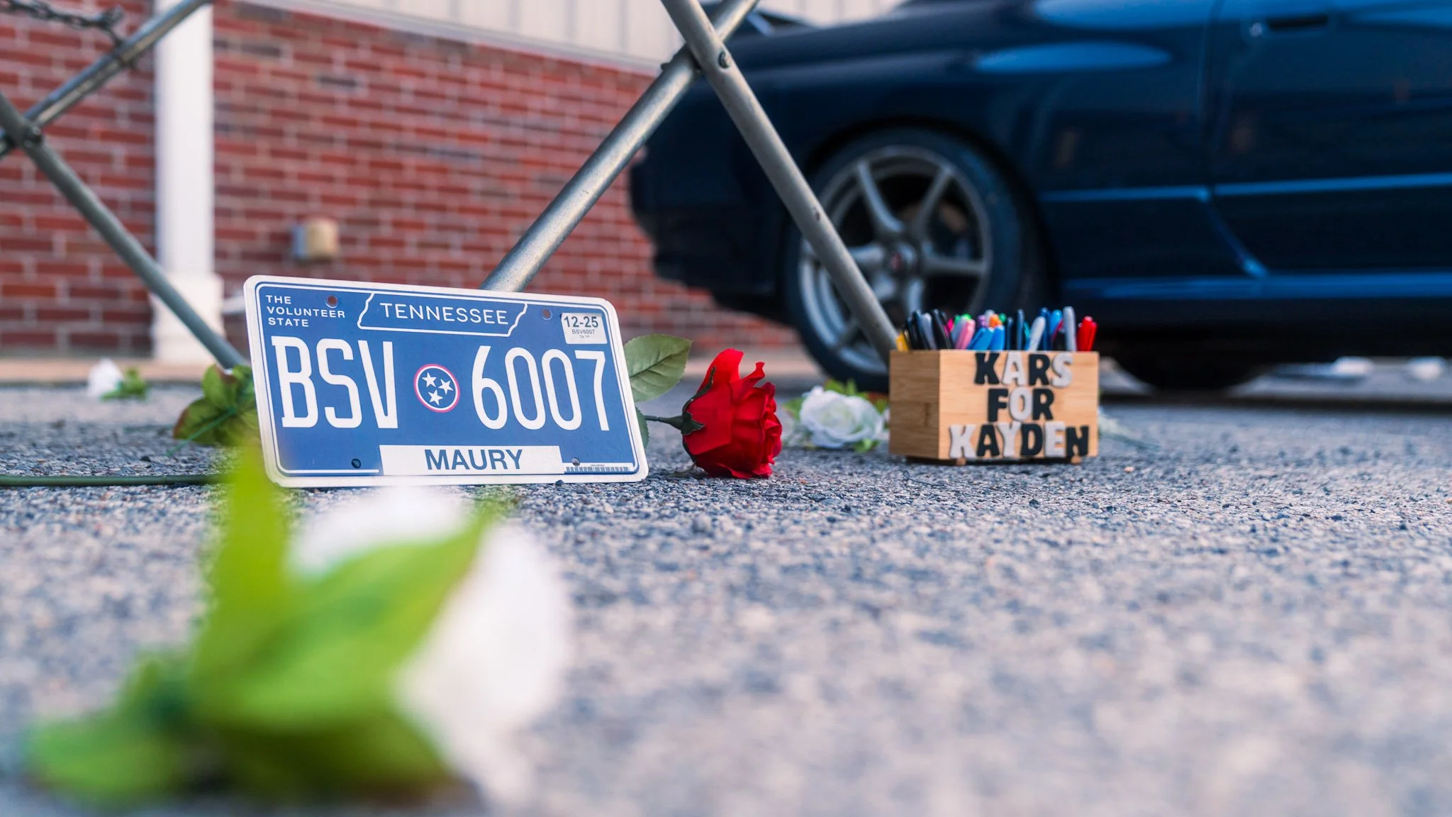 Memorial scene on pavement with a Tennessee license plate reading 'MAURY', surrounded by white and red flowers, a small wooden box labeled 'KARS FOR KAYDEN' filled with colorful markers, a black car, and a brick building in the background.