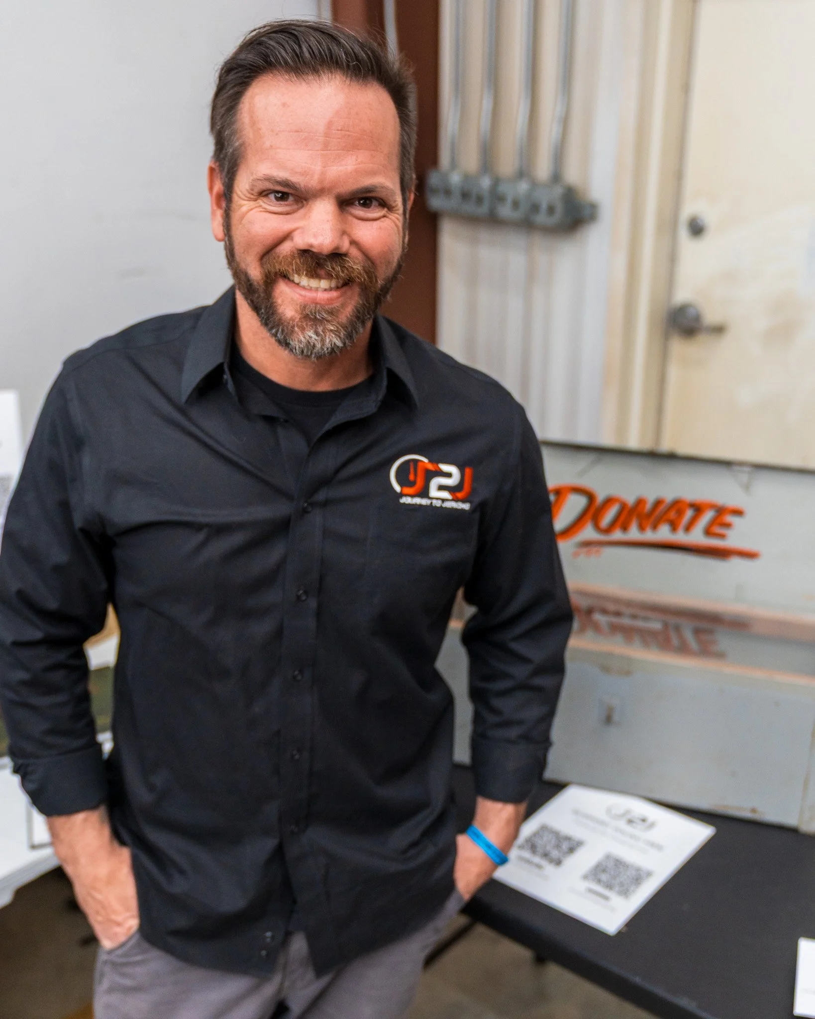 A man with a beard smiling, wearing a black button-up shirt with a logo on the chest that displays 'JC' and 'Jeremy Jansen', standing in front of a sign that says 'DONATE' and some QR codes on a table.