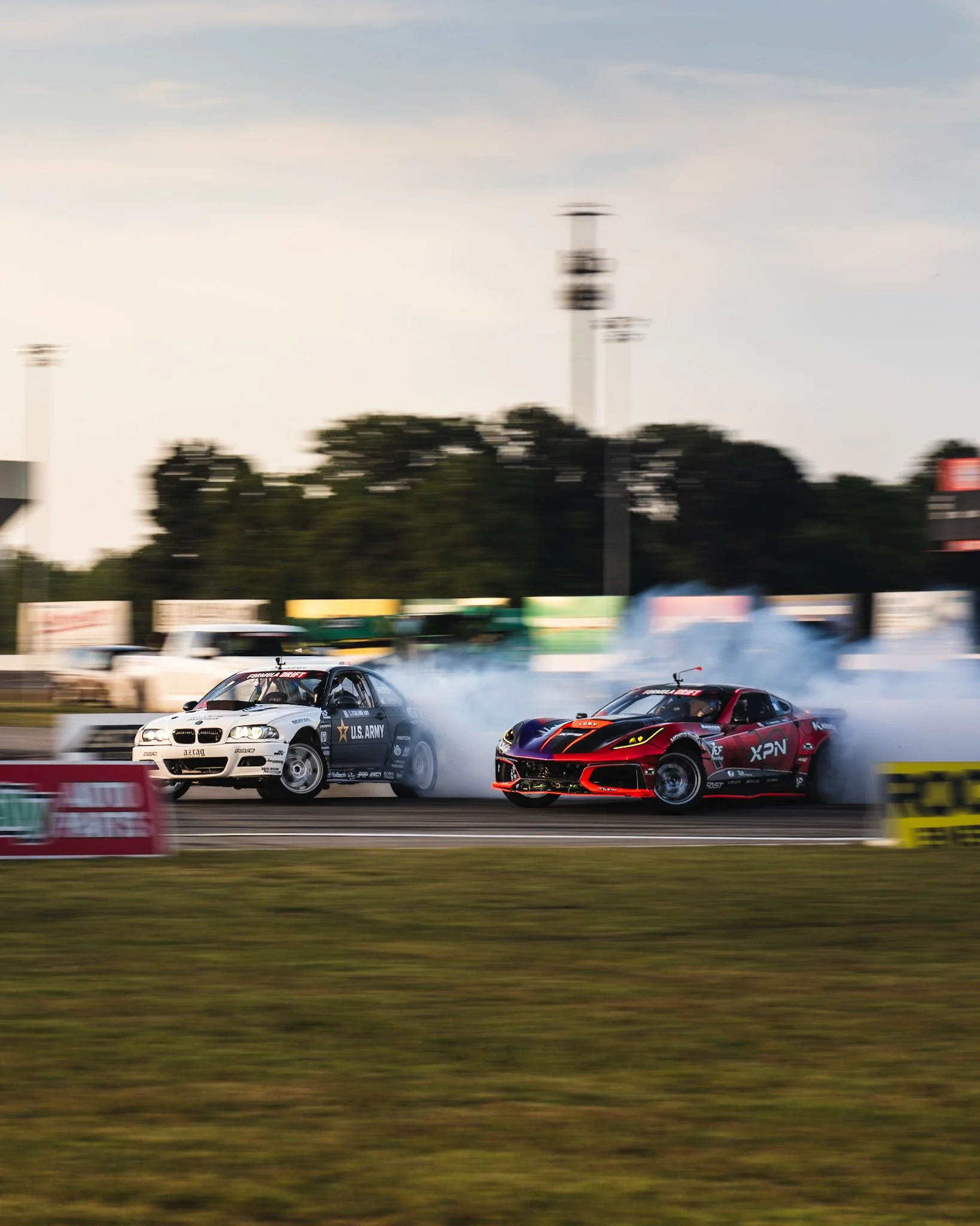 Two race cars drifting and creating smoke on a race track, with trees, advertisements, and a cloudy sky in the background. Formula Drift Indy