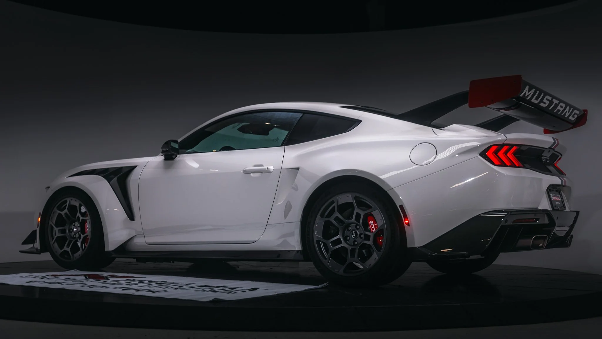 White Ford Mustang GT with racing stripes, large rear wing, and black wheels on display against a dark background.