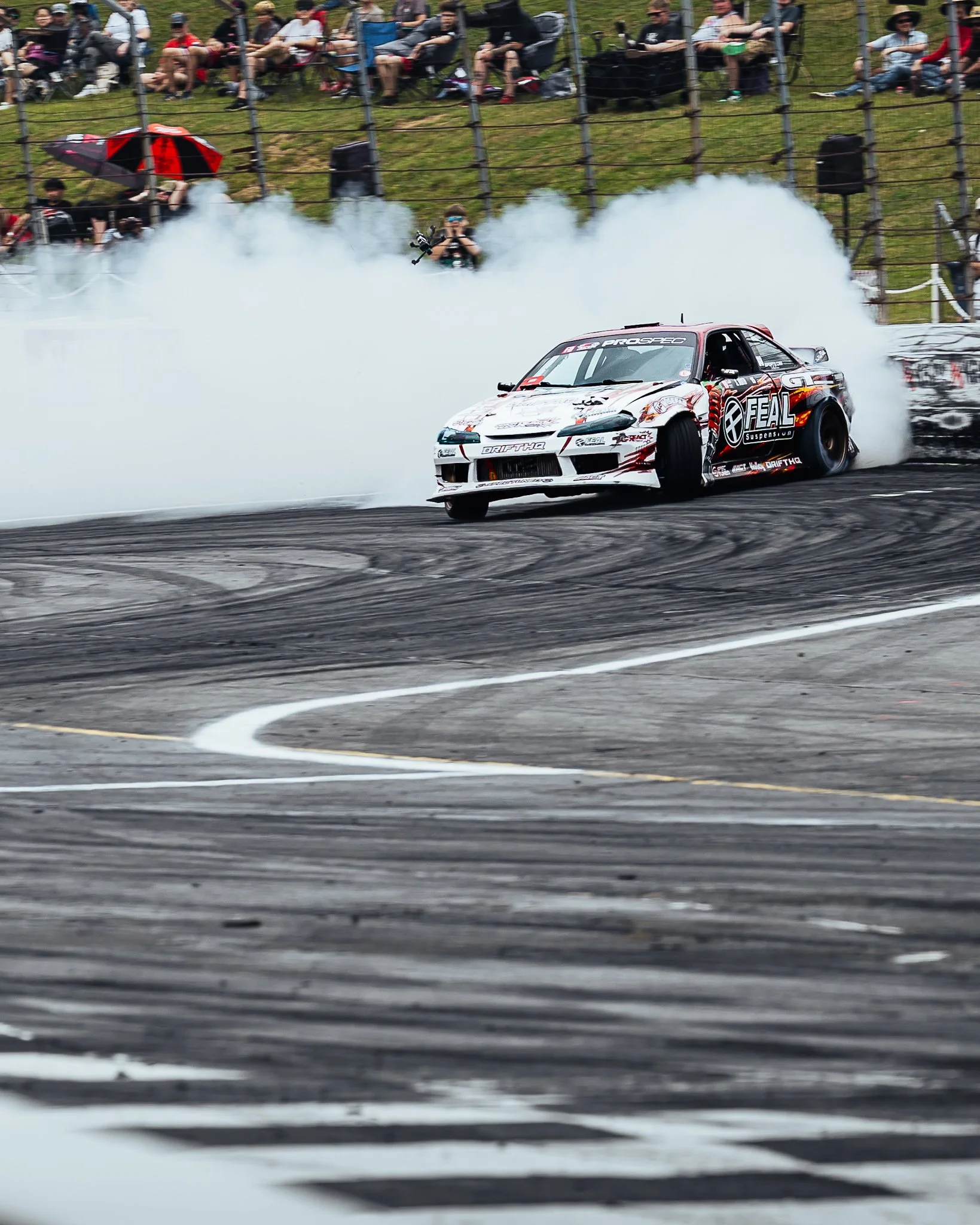 A race car drifting on a racetrack, producing smoke, with spectators watching from the stands.