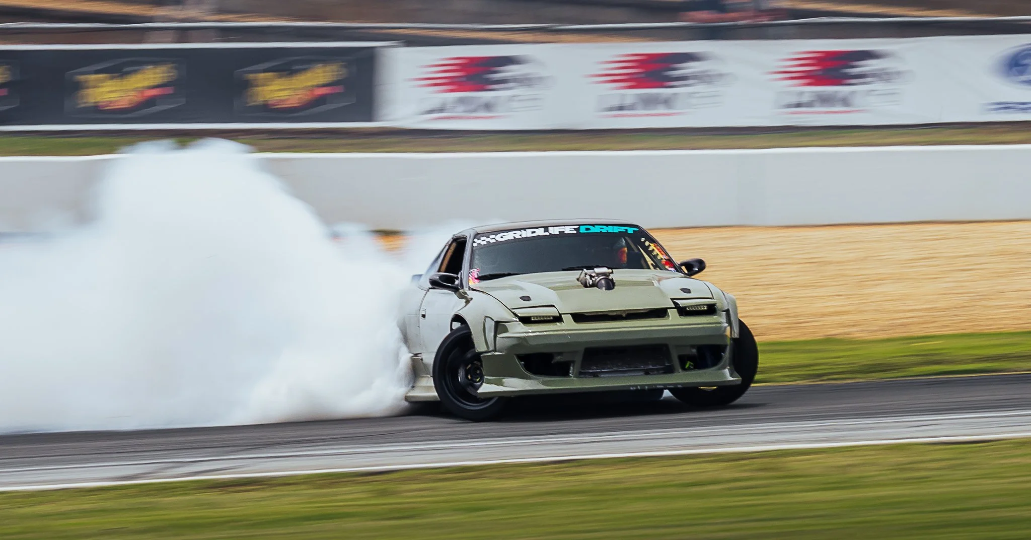 A silver sports car drifting on a racetrack, producing smoke from the tires with blurred colorful advertisement banners in the background.