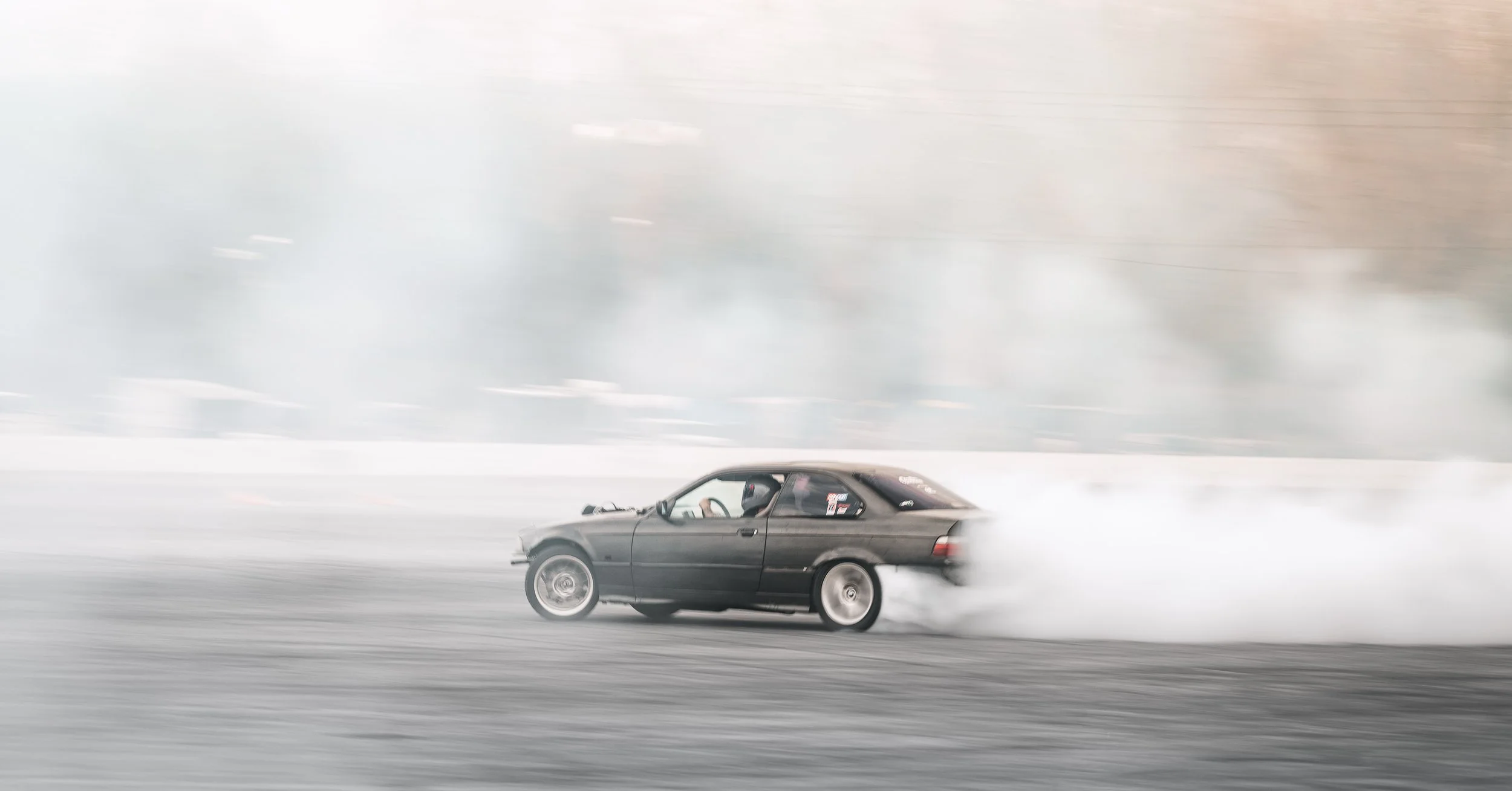 A black car drifting on a snowy surface, creating a spray of snow behind it.