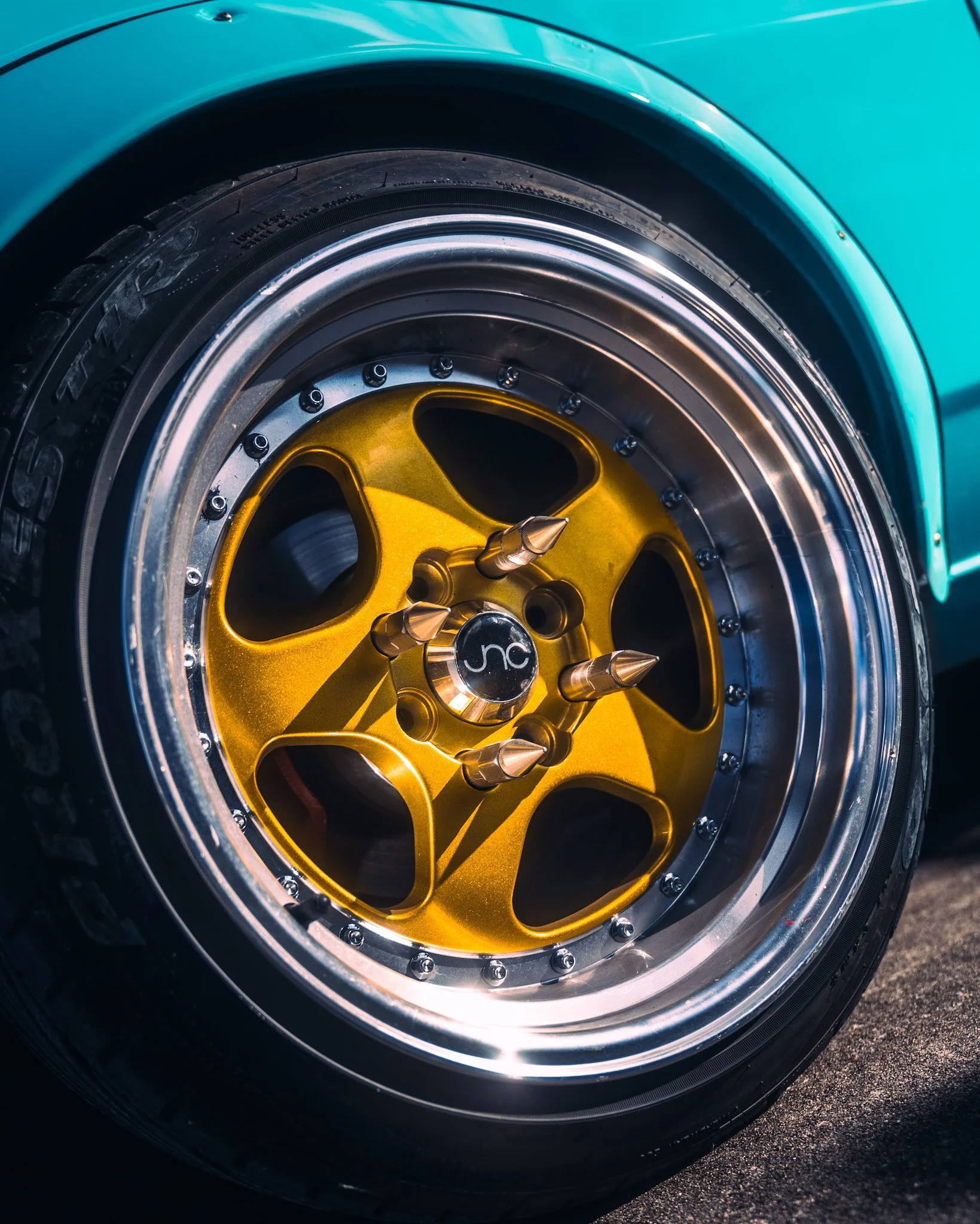 Close-up of a car wheel with a yellow rim, black tire, and a teal-colored body in the background.