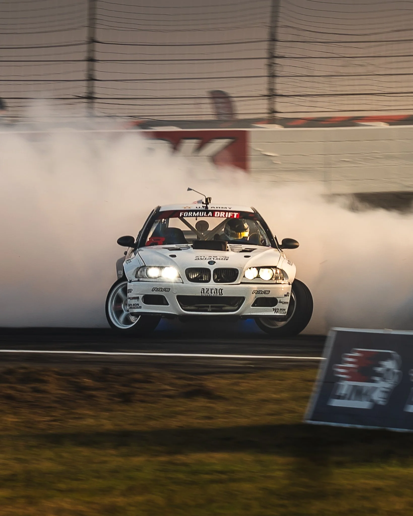 A white Formula Drift race car with various sponsor stickers on it, driving on a track and kicking up smoke as it performs a drift. The background includes a fence and a sunset sky. Connor Osully Formula Drift Indy