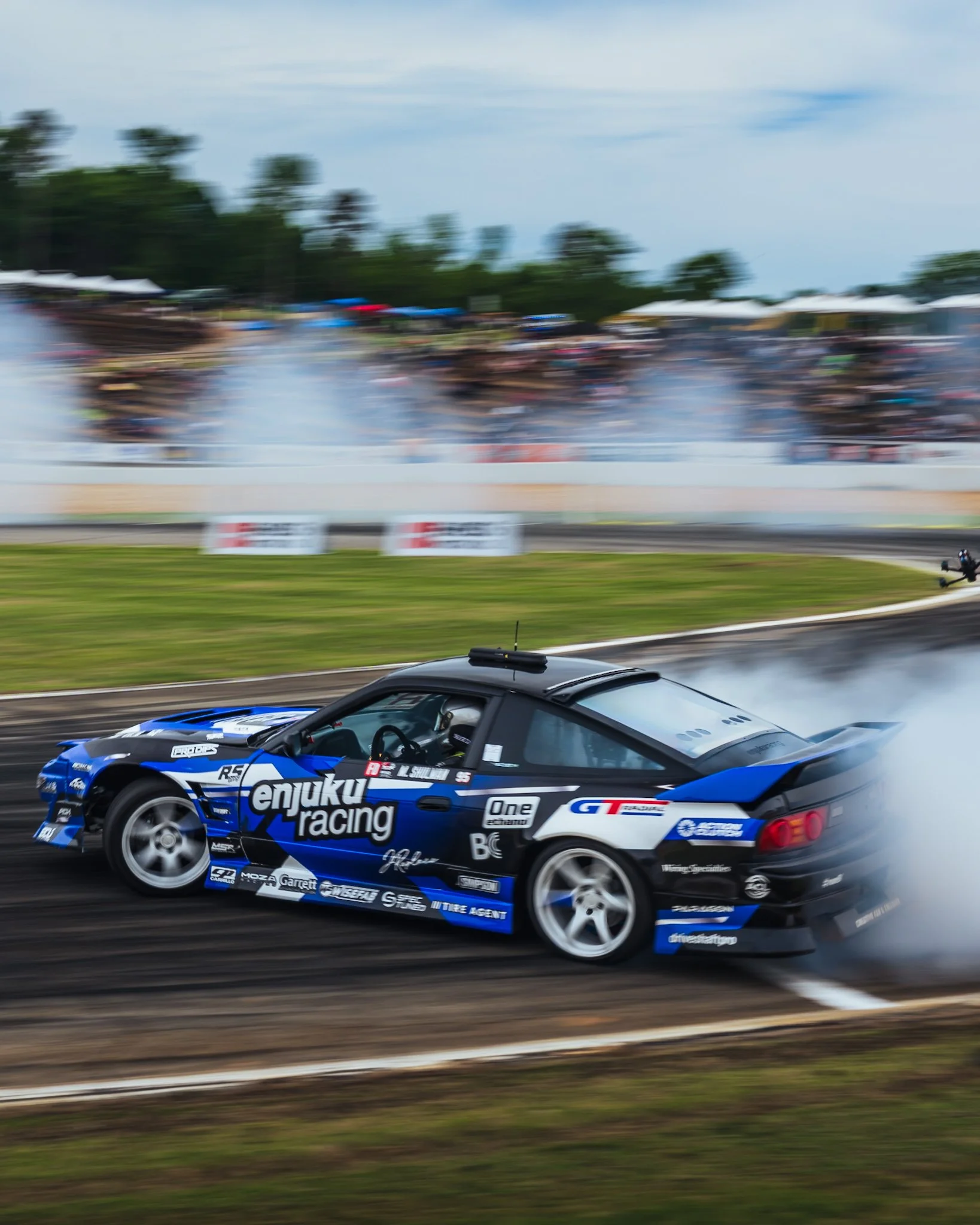 A racing car with blue and black livery passing by on a race track, with spectators and trees blurred in the background.