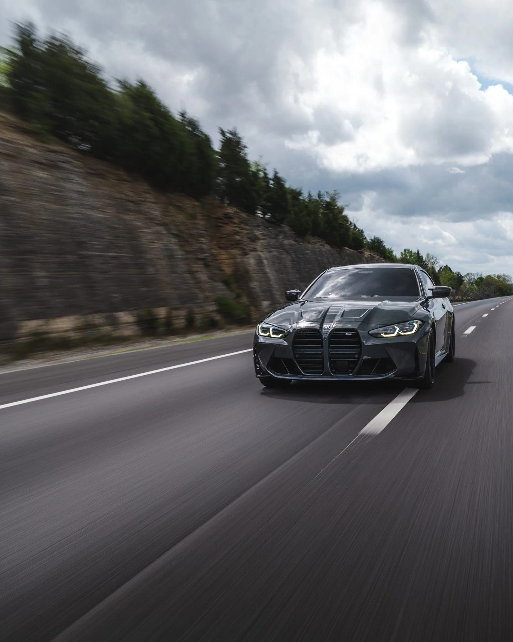 A black sports car driving on a highway with a rocky hill and trees in the background under cloudy skies.