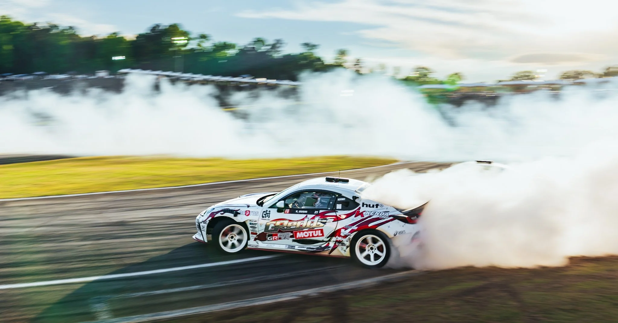 A white racing car with red and black decals speeds on a track, creating a trail of smoke behind it during a drift or race, with a crowd of spectators and trees in the background.