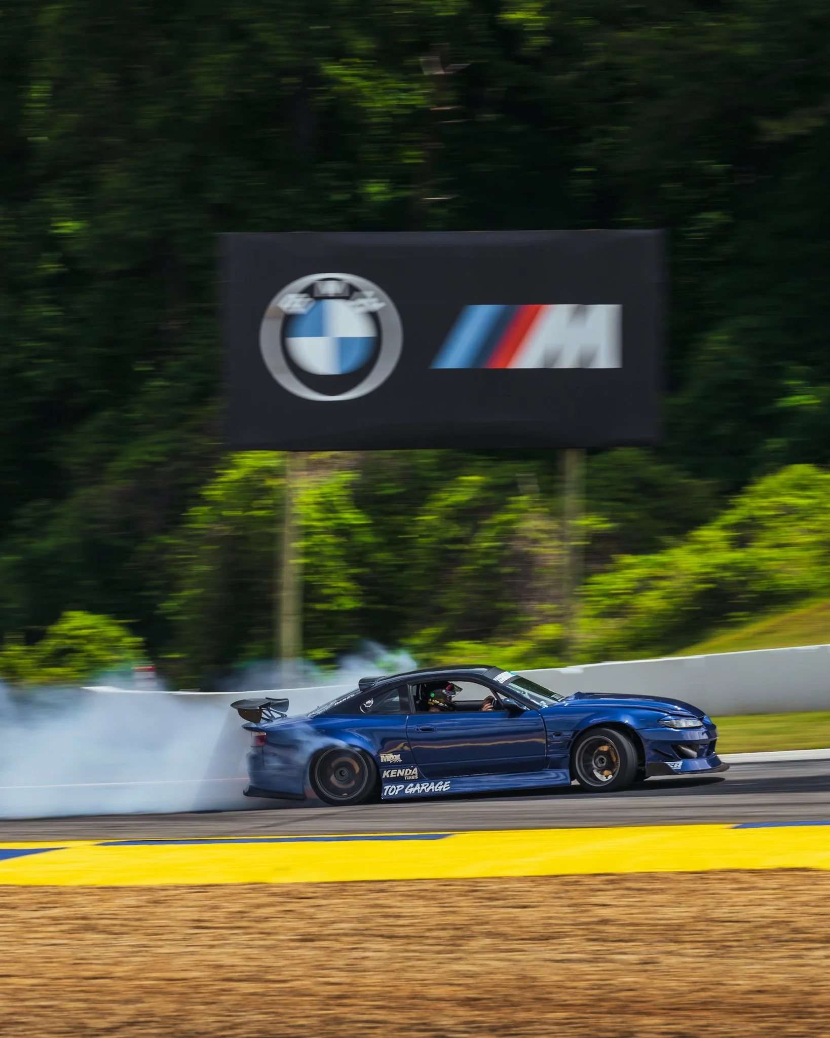 A blue racing car drifting on a race track, producing smoke from the tires, with a driver wearing a helmet inside. There is a black banner in the background with the BMW logo and other racing-related logos, and green foliage beyond the banner.