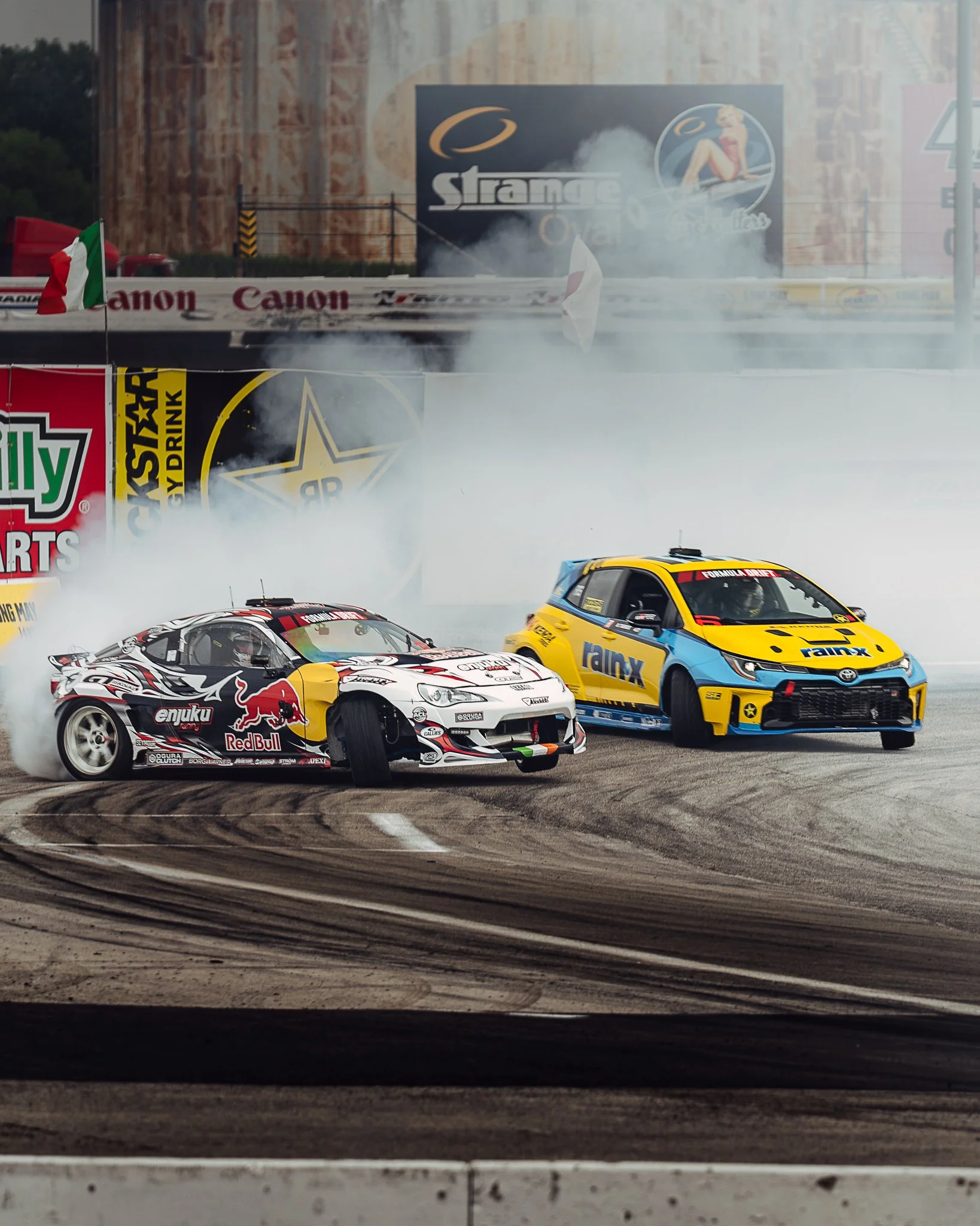 Two race cars drifting on a track with tire smoke, with advertising banners and large signage in the background, and an Italian flag on the left side. Formula Drift Indy